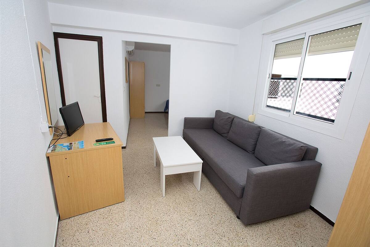 Una habitación pequeña y poco amueblada, con un escritorio de madera, una mesa blanca y un sofá gris. Cuenta con una ventana con persianas, una puerta que da a otra habitación y un espejo en la pared.