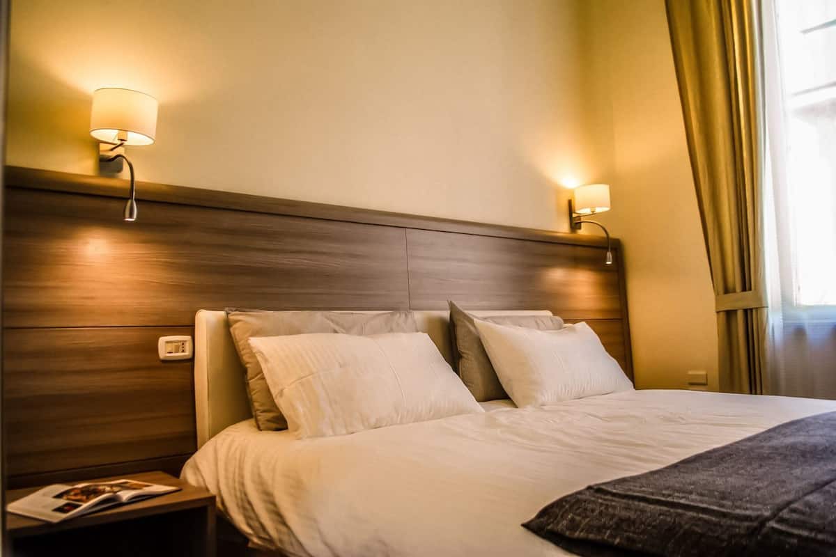 Habitación de hotel con cabecera de madera, dos lámparas de noche, cama con ropa blanca y mesita de noche con una revista.