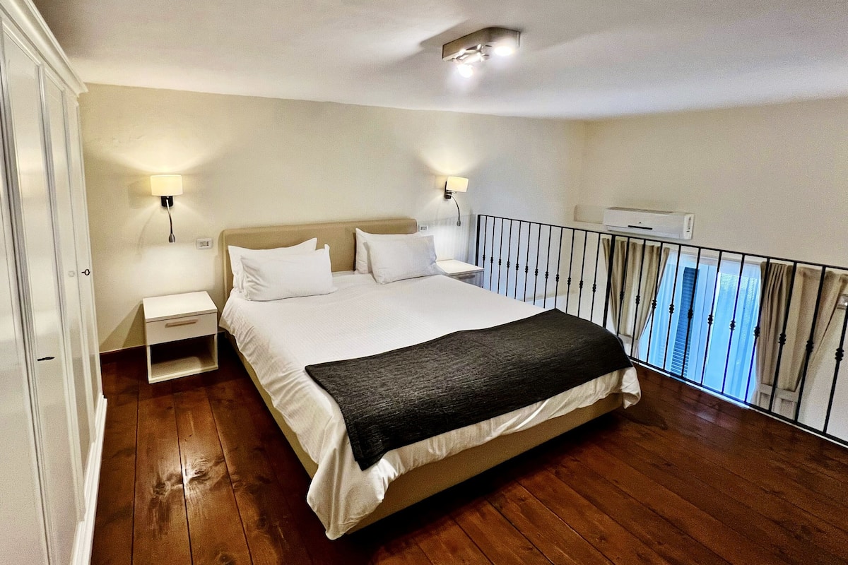 Un dormitorio con una cama grande, dos mesitas de noche, una mesita auxiliar, una lámpara en el techo y un balcón con barandilla.