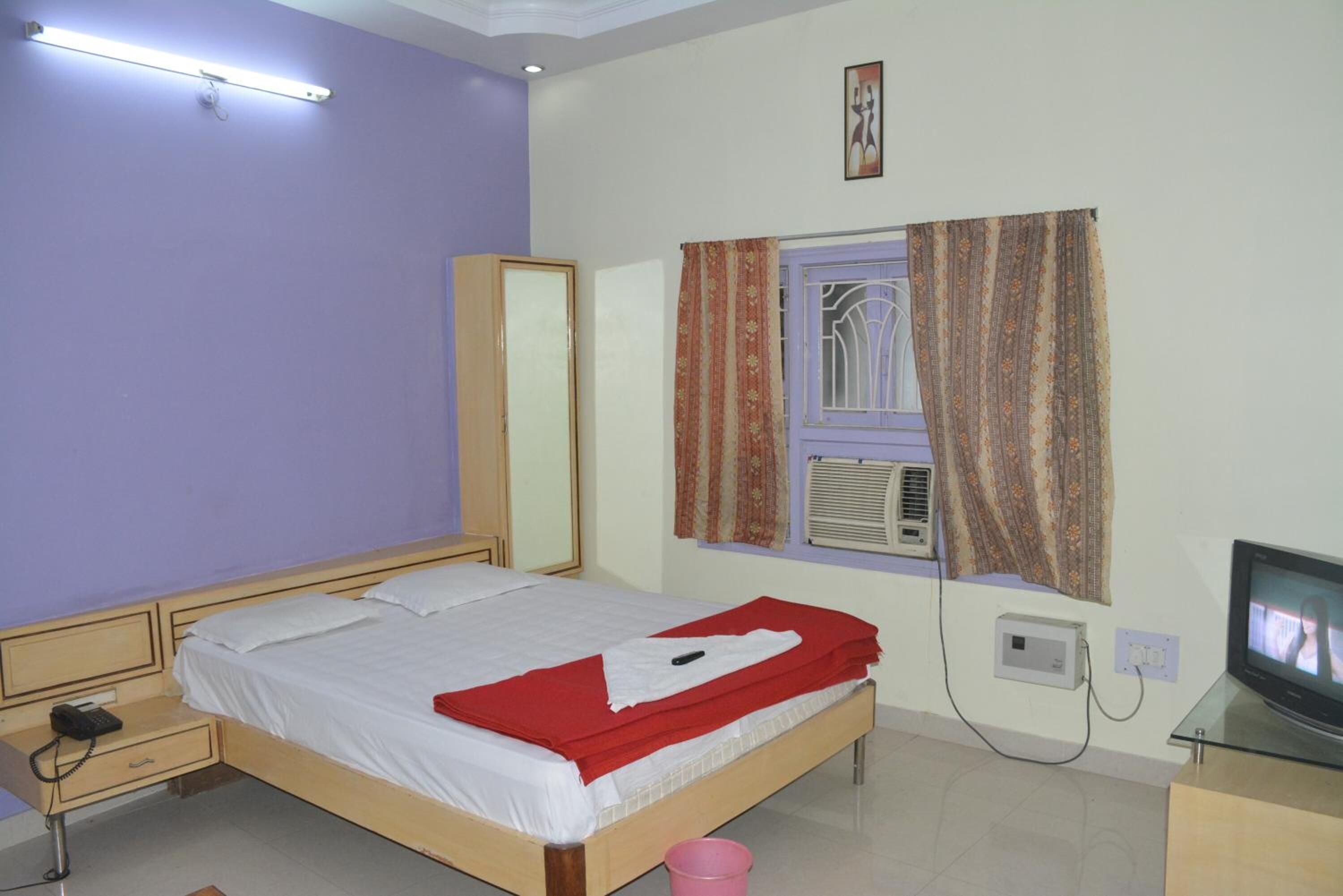 A bedroom with a bed, a bedside table, a mirror, a window with curtains, an air conditioning unit, and a television.