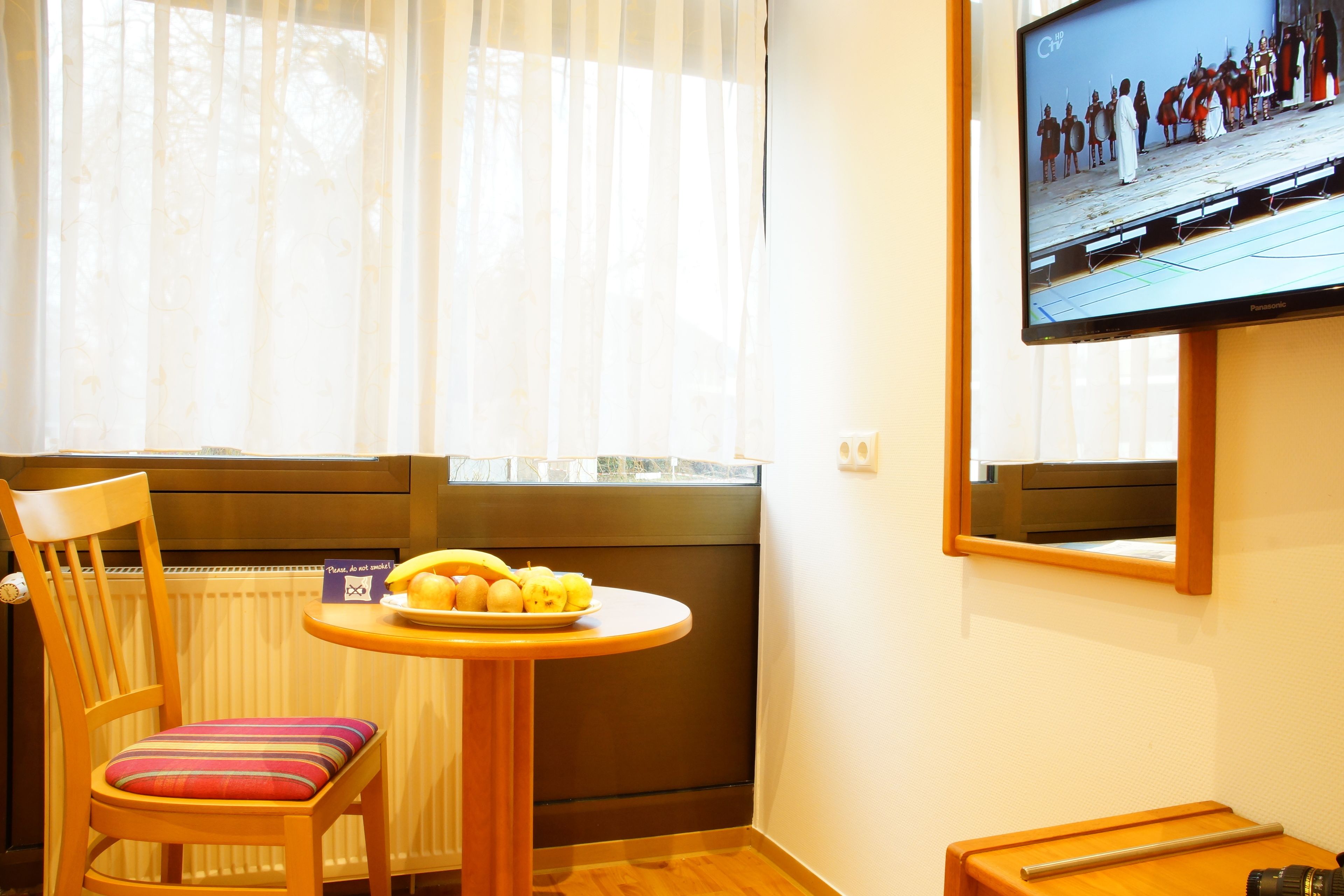 A room with a wooden chair, a small round table with a plate of fruit, a television mounted on the wall, and a window with sheer curtains.