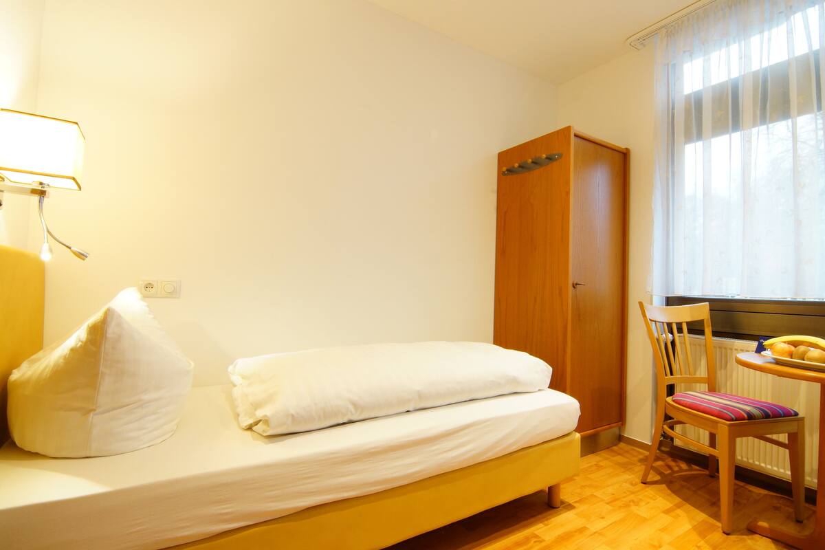 A single bed with white bedding, a wooden wardrobe, a chair with a striped cushion, and a table with fruits.
