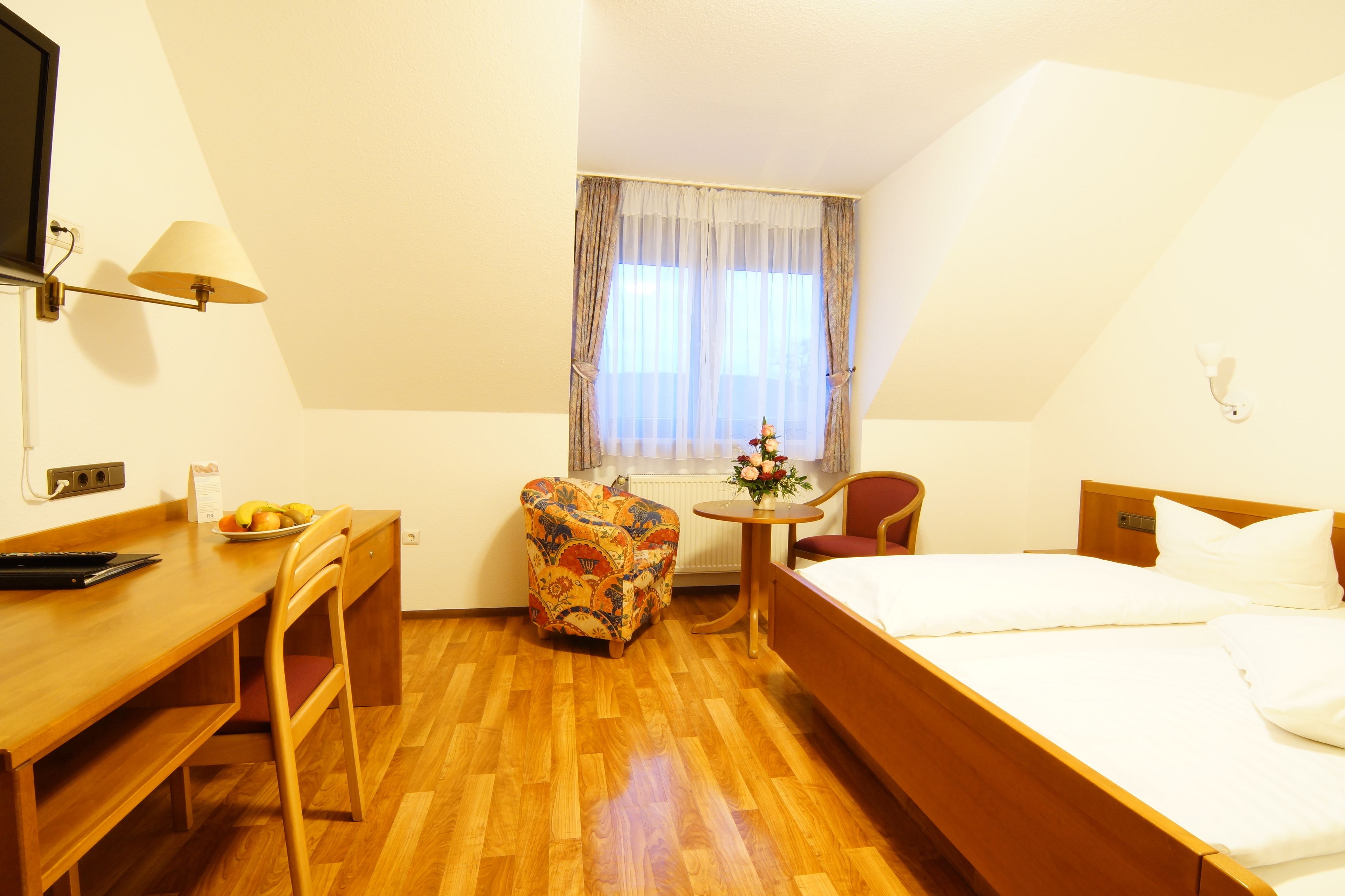 A hotel room with a wooden bed, a desk with a chair, a small table with a chair, and a window with curtains.
