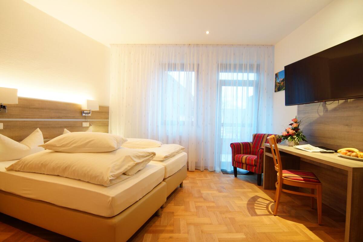 A hotel room with a bed, a desk, a chair, a flat-screen TV, and a window with sheer curtains.