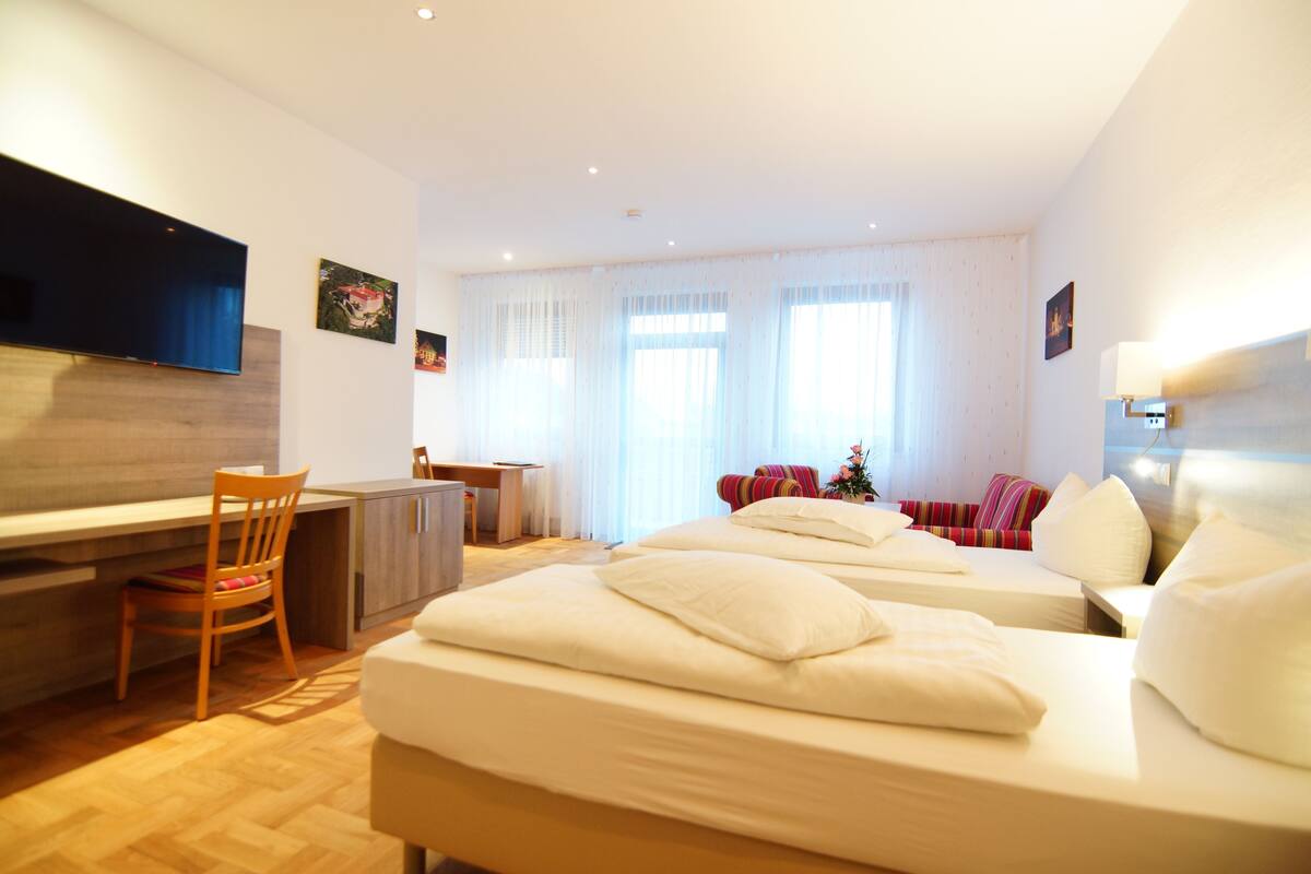 A hotel room with two beds, a desk, a chair, a TV, and a large window with curtains.