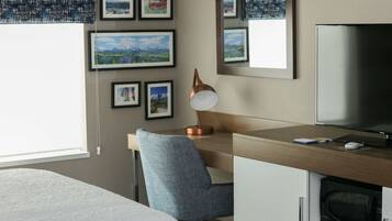 A hotel room with a bed, a desk with a lamp, a TV, and framed pictures on the wall.