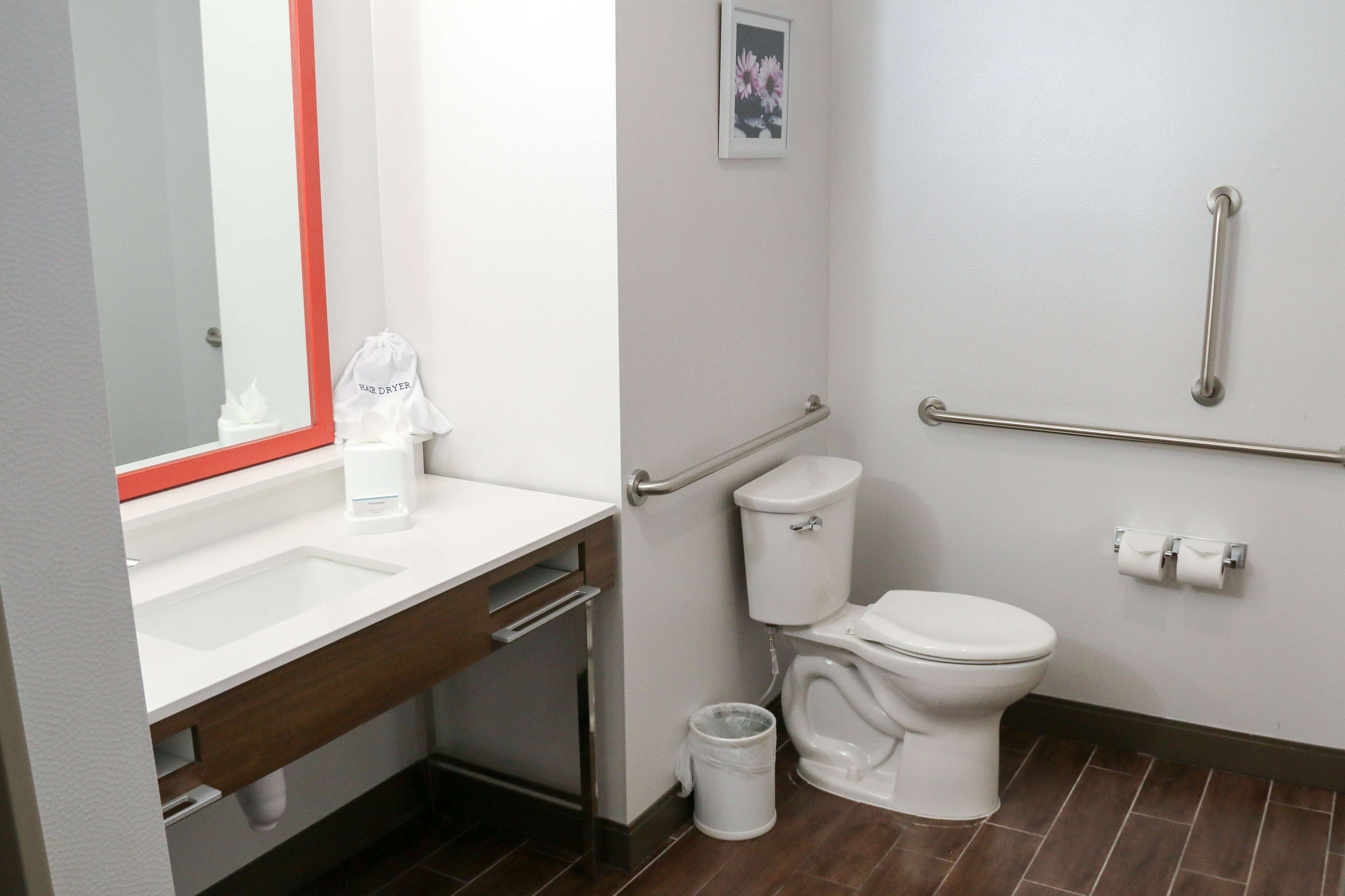 A modern bathroom with a white sink, mirror, and toilet. There is a hand dryer and a trash can.