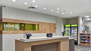 A hotel reception desk with a modern design, a digital check-in station, and a refrigerator displaying 'cold' and 'rearily cold' beverages. - Home2 Suites by Hilton McKinney (McKinney)