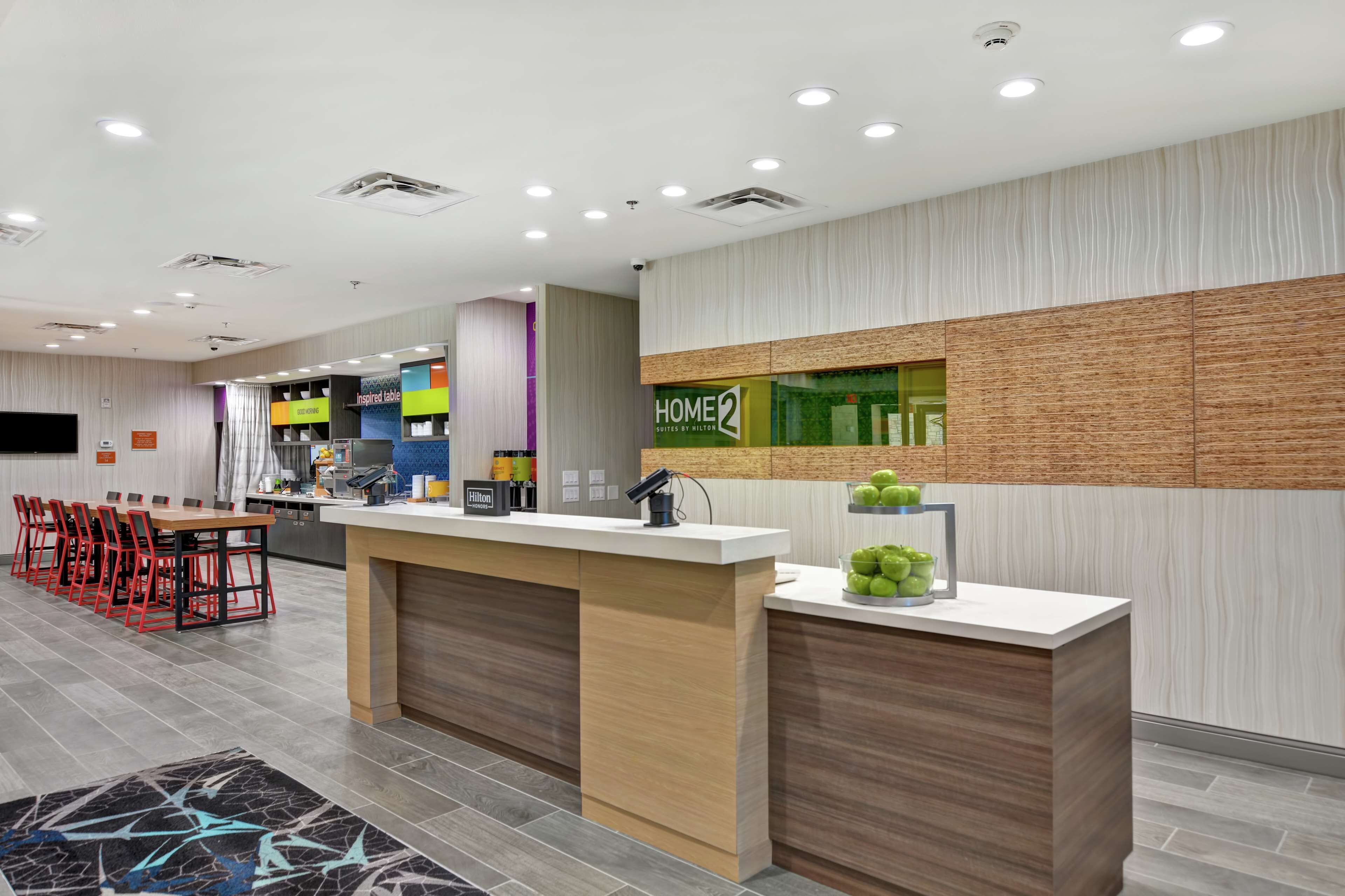 A modern hotel lobby with a reception desk, a seating area with red chairs, and a kitchen in the background.