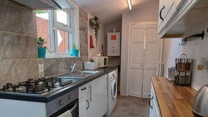 Een praktische keuken met een gasfornuis, een inbouwoven, een magnetron en een wasmachine. Er is een raam met potplanten, een witte deur en een aanrecht met potten.
