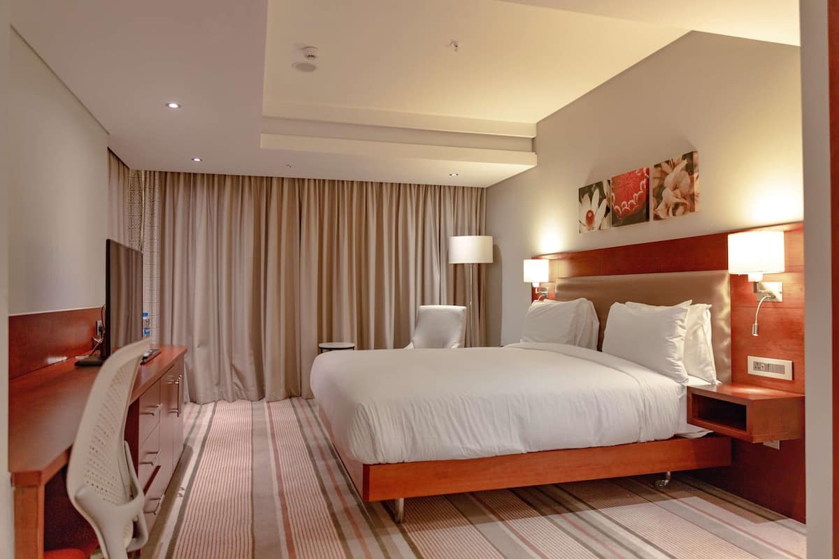 A hotel room with a bed, desk, chair, lamp, and artwork on the wall.