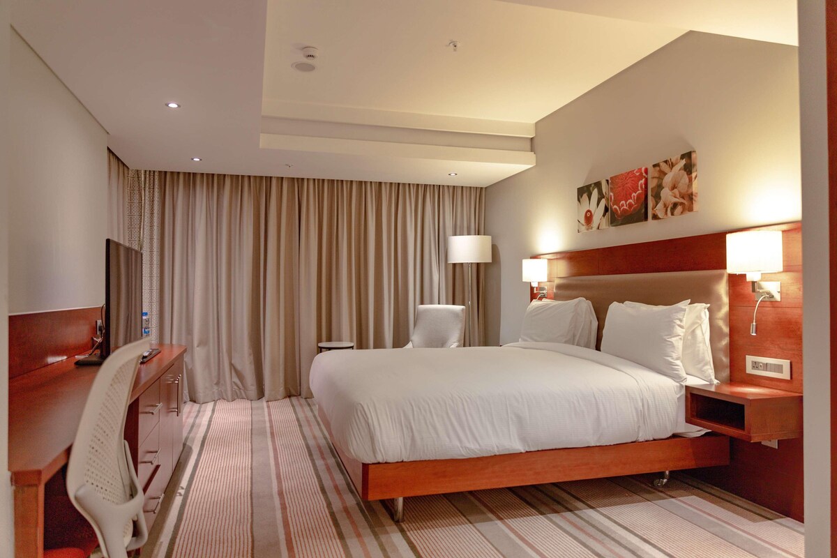 A hotel room with a bed, desk, chair, lamp, and artwork on the wall.