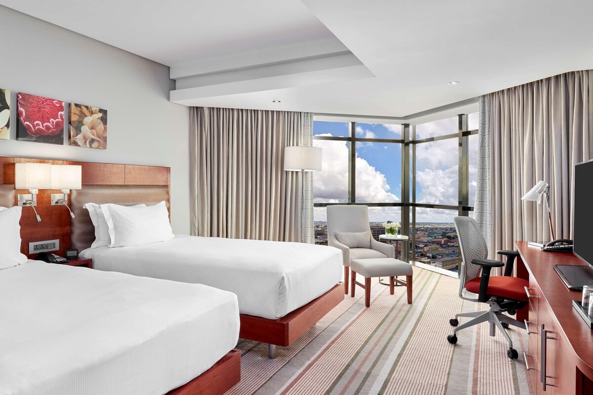 Habitación de hotel con dos camas, un escritorio con ordenador, una silla, una lámpara y vistas de la ciudad a través de amplias ventanas.