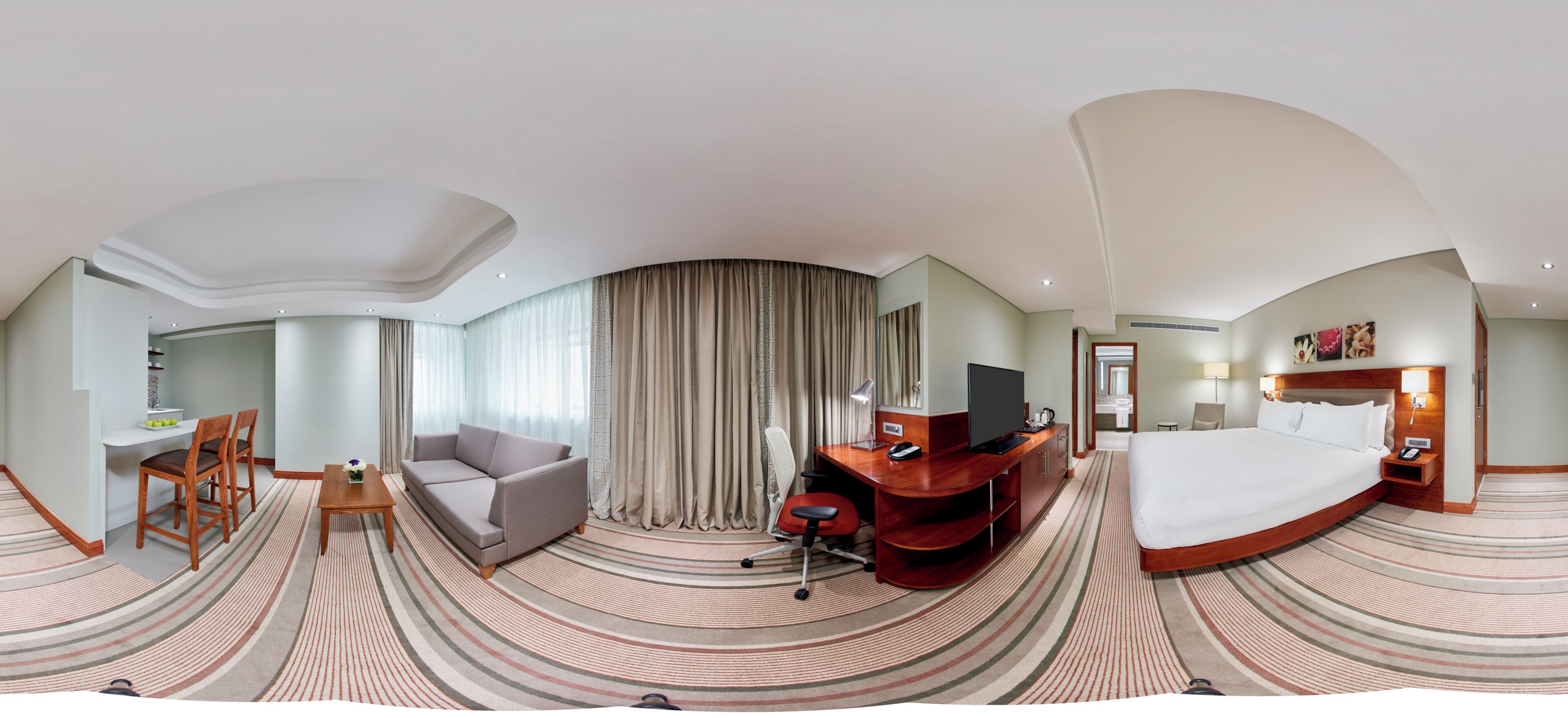 A panoramic hotel room with a curved ceiling, a bed, a desk with a chair, a sofa, and a TV.