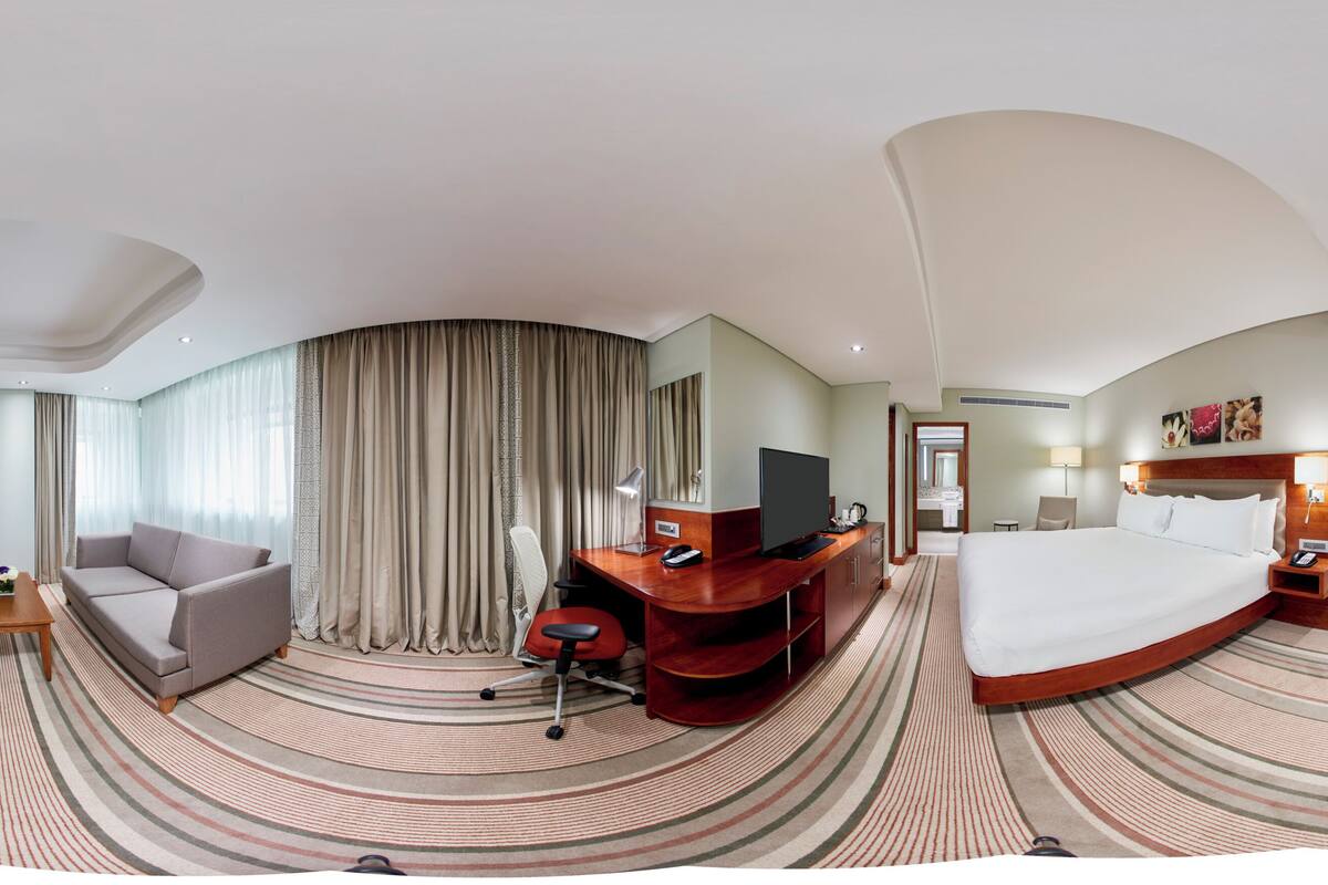 A panoramic hotel room with a curved ceiling, a bed, a desk with a chair, a sofa, and a TV.
