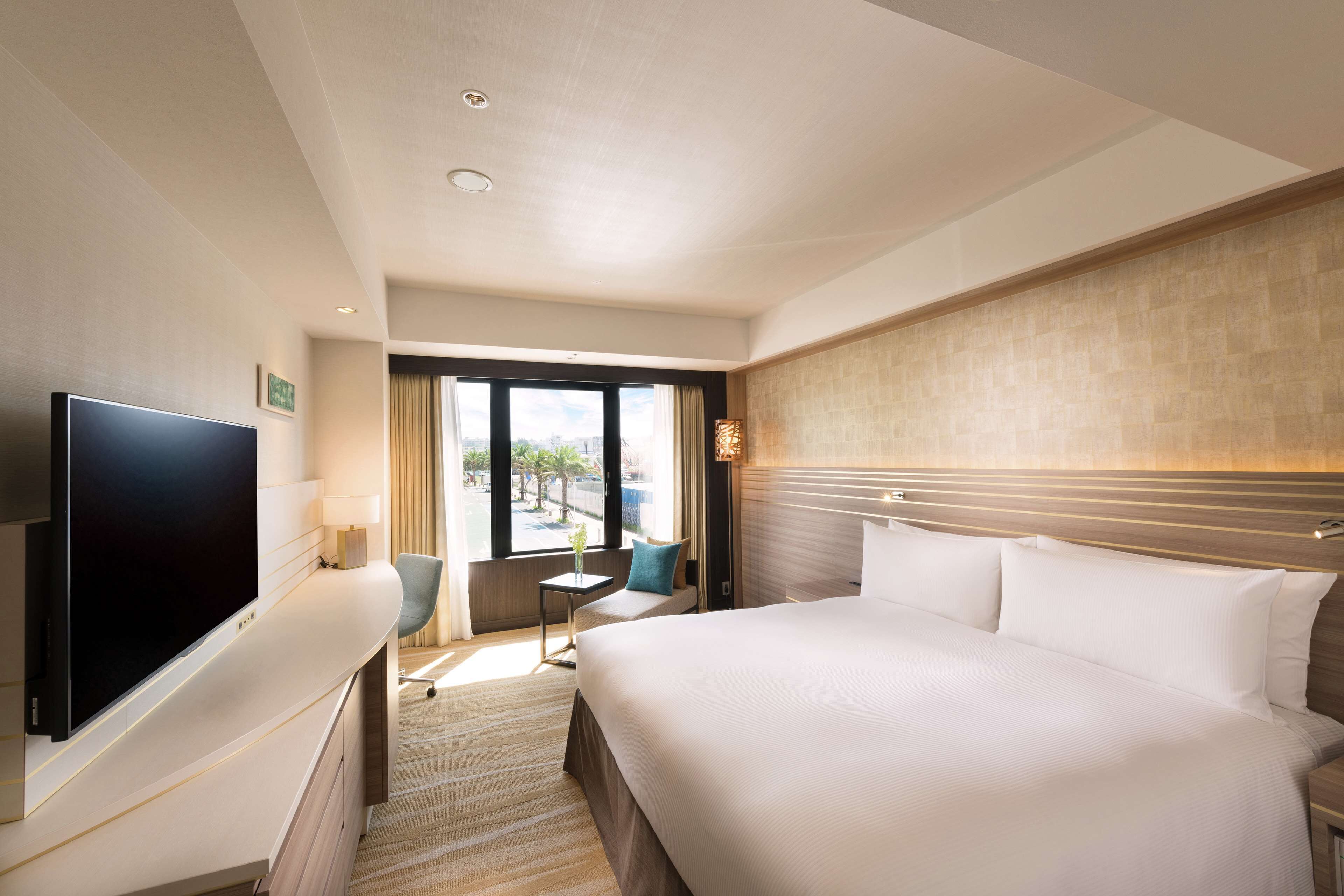A hotel room with a large bed, a flat-screen TV, a small table with a lamp, and a view of the outdoors through a large window.