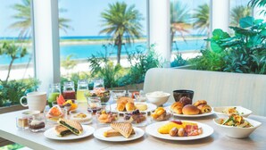 Un bufé de desayuno con diversas opciones de comida y bebidas, un ventanal con vista a la playa y palmeras.