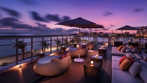 Una terraza en la azotea con zonas de estar, mesas y vistas del horizonte urbano al atardecer.