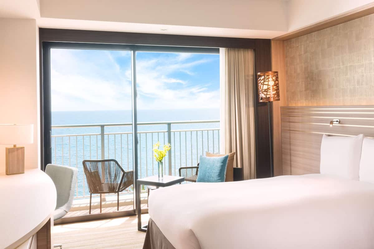 A hotel room with a large balcony, a bed, a desk, and a chair.