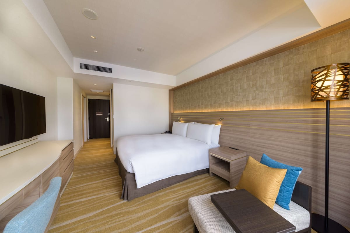 A modern hotel room with a large bed, bedside tables, a flat-screen TV, and a seating area with a bench and cushions.