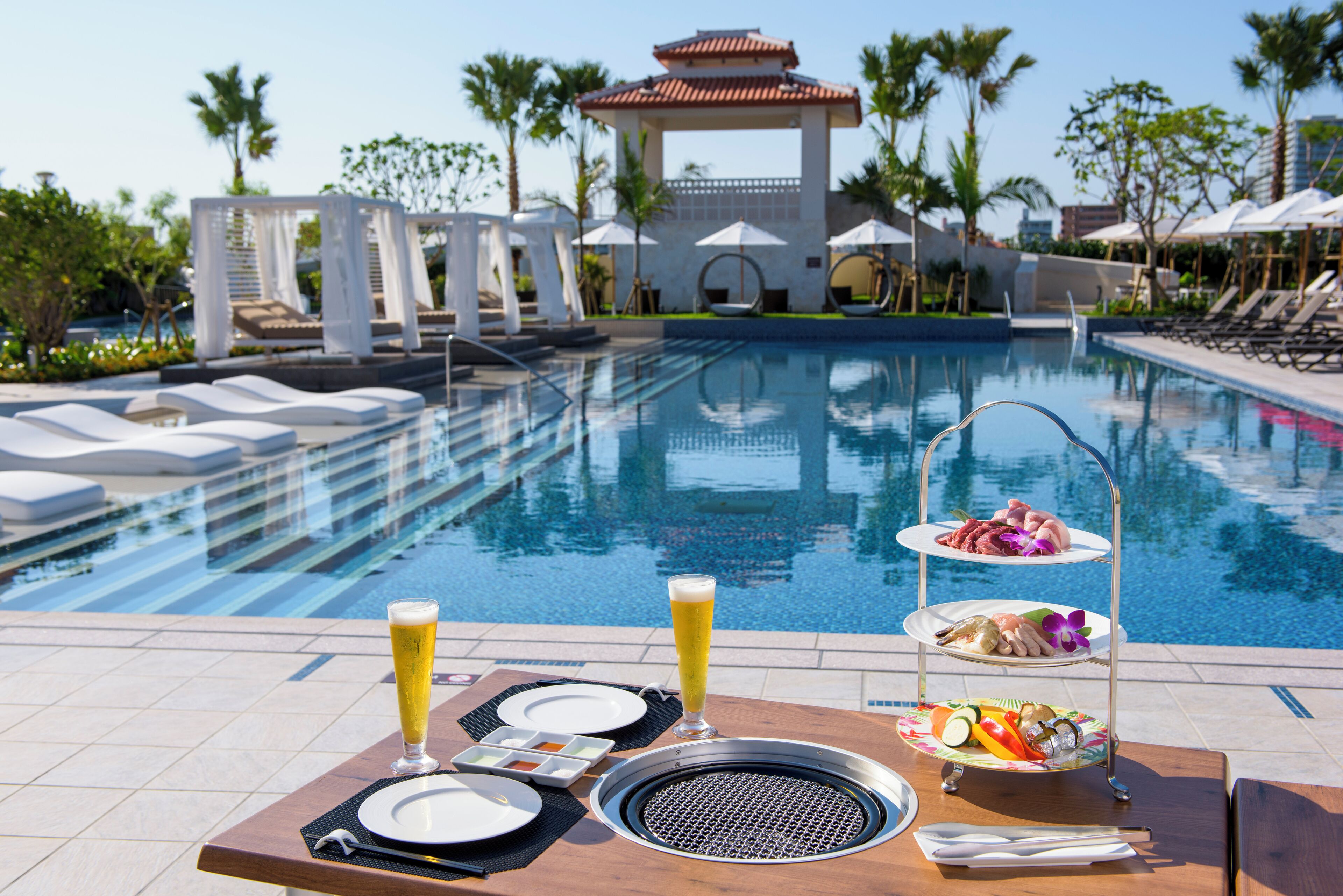 A poolside dining setup with a table, plates, cutlery, and a three-tiered food and drink stand.