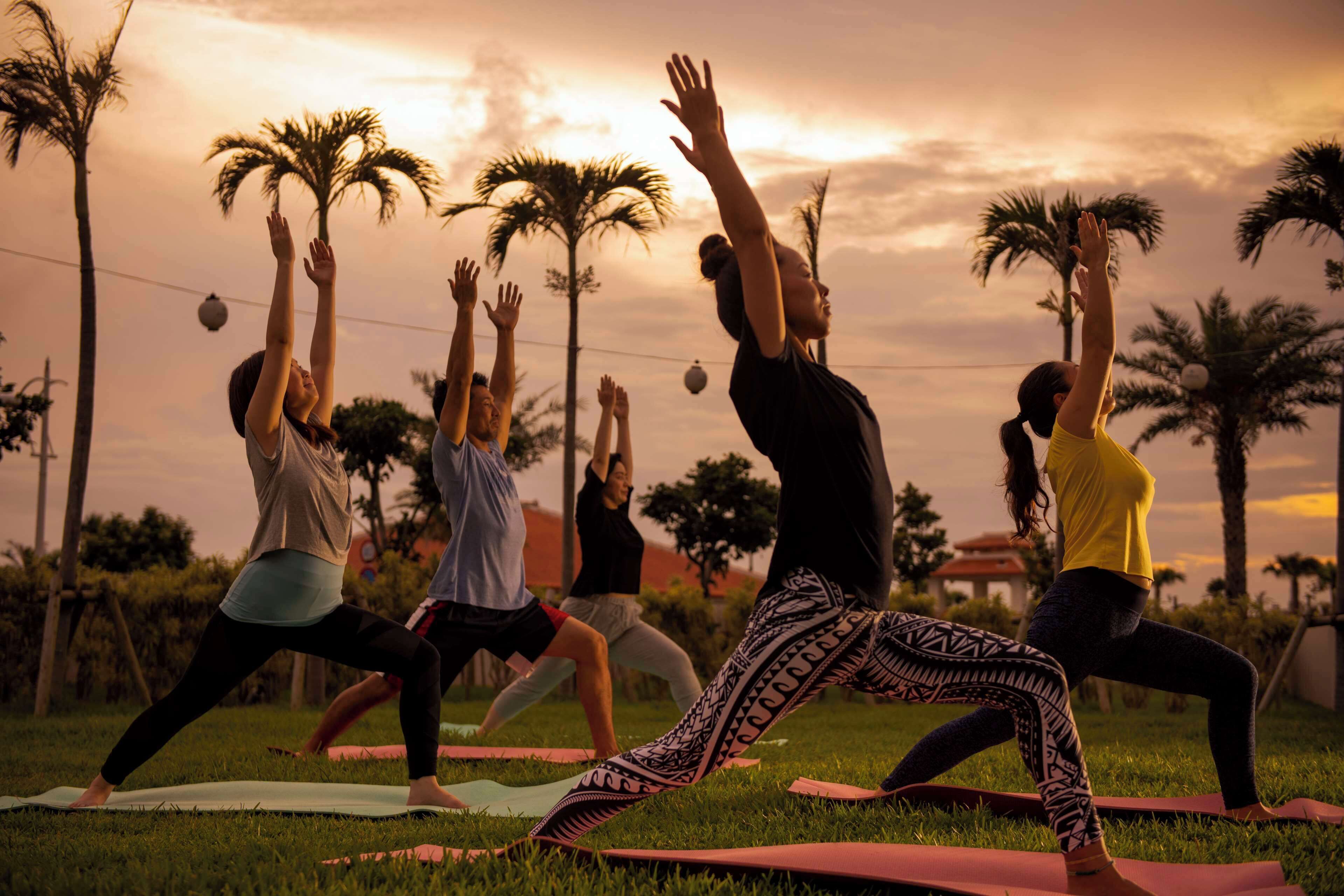 A group of people performing yoga on mats in an outdoor setting with palm trees and a sunset in the background.