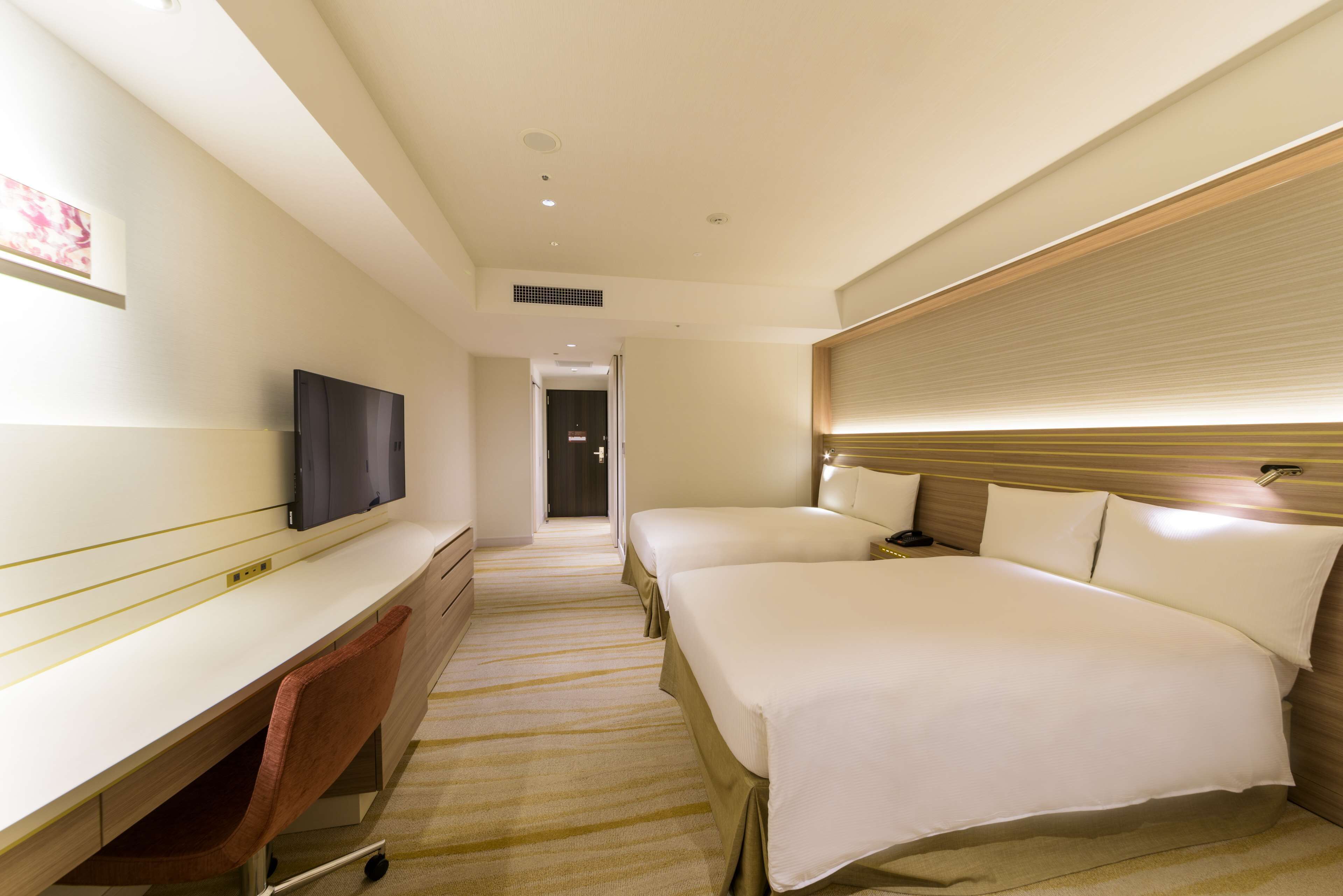 A hotel room with a desk, two beds, a TV, and a headboard with built-in lighting.