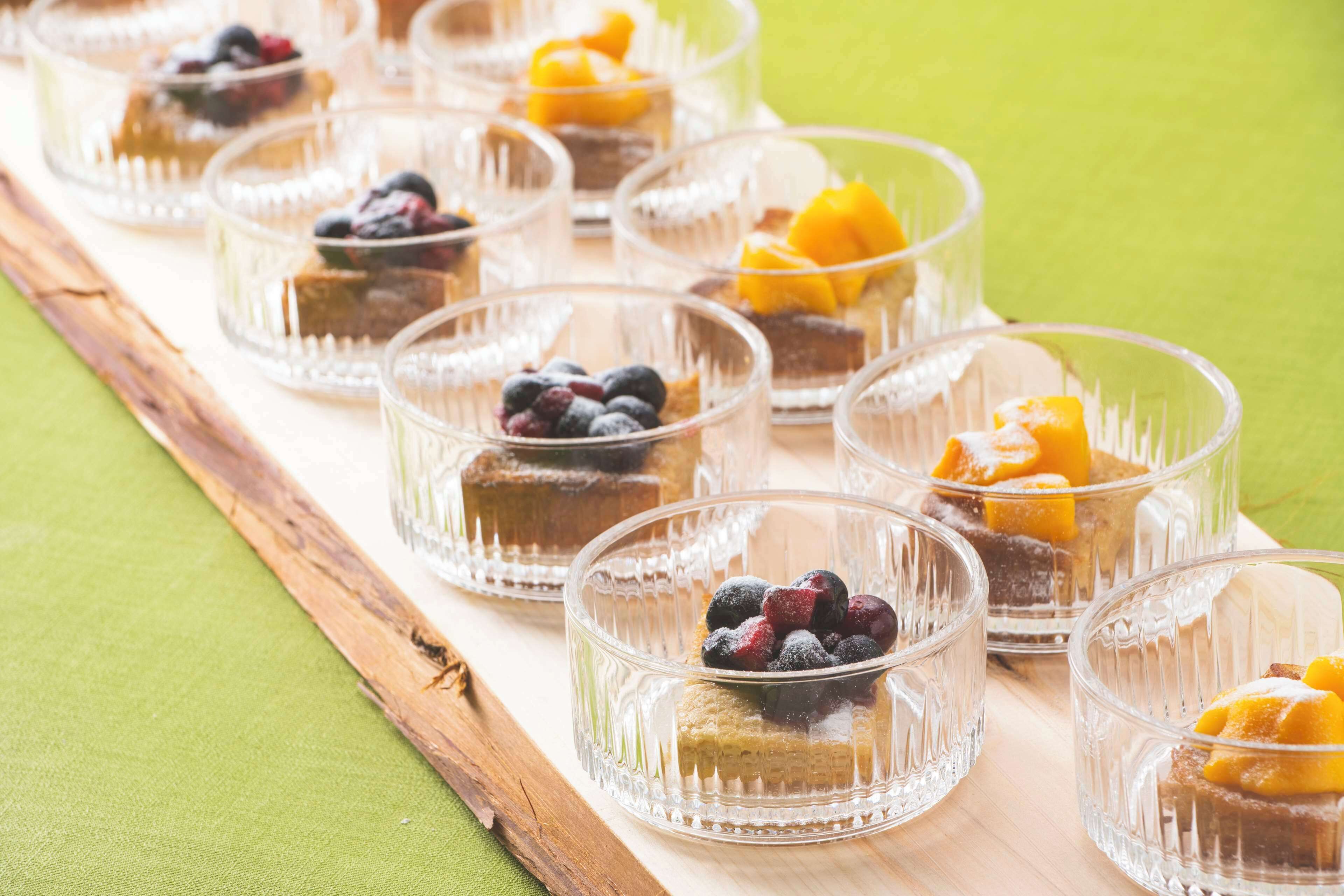 A series of clear glass dessert cups arranged on a wooden tray, each topped with a slice of cake and various fruits.