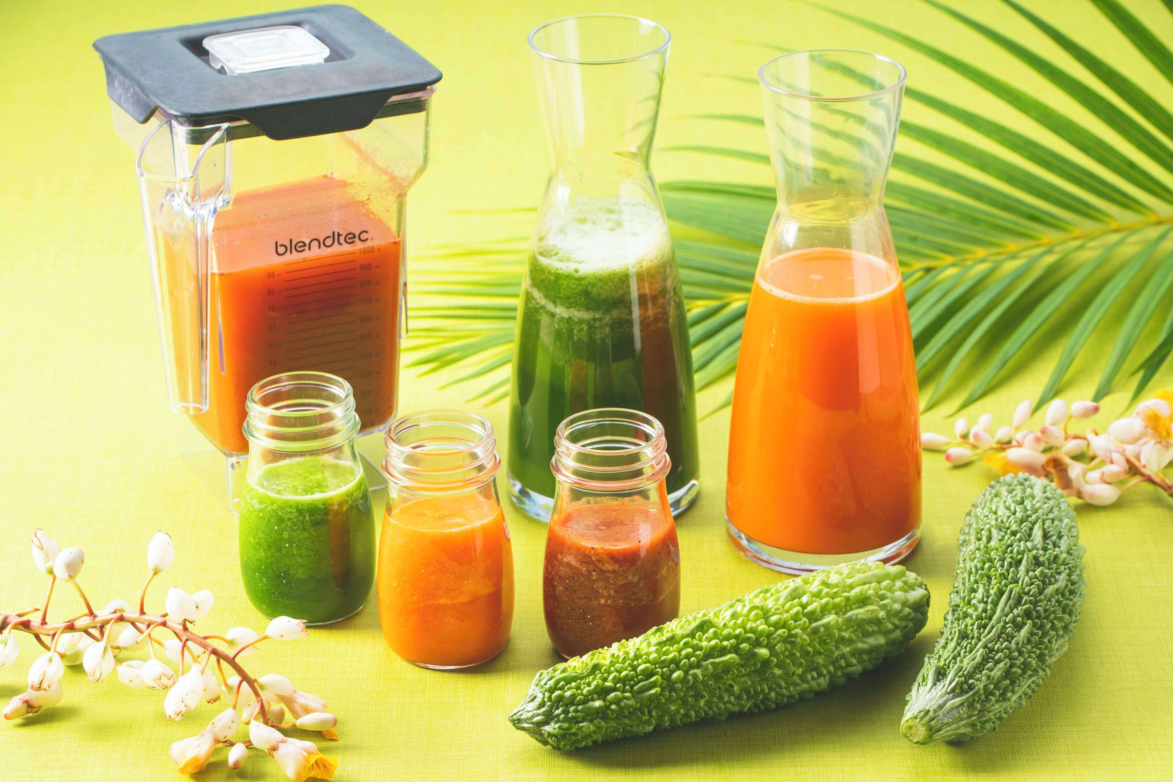 A variety of glass containers with different colored liquids, a blender, and two bitter gourds on a green surface.