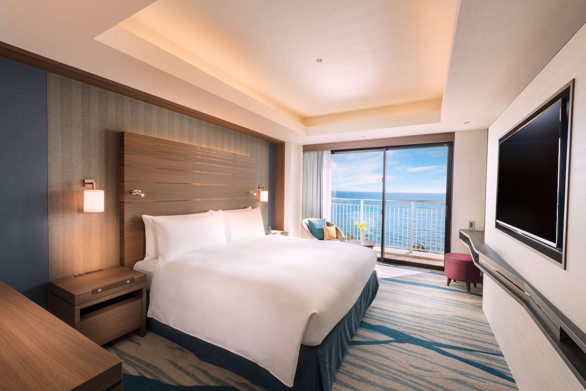 A modern hotel room with a large bed, a TV, a balcony with a view of the ocean, and a nightstand with a lamp.