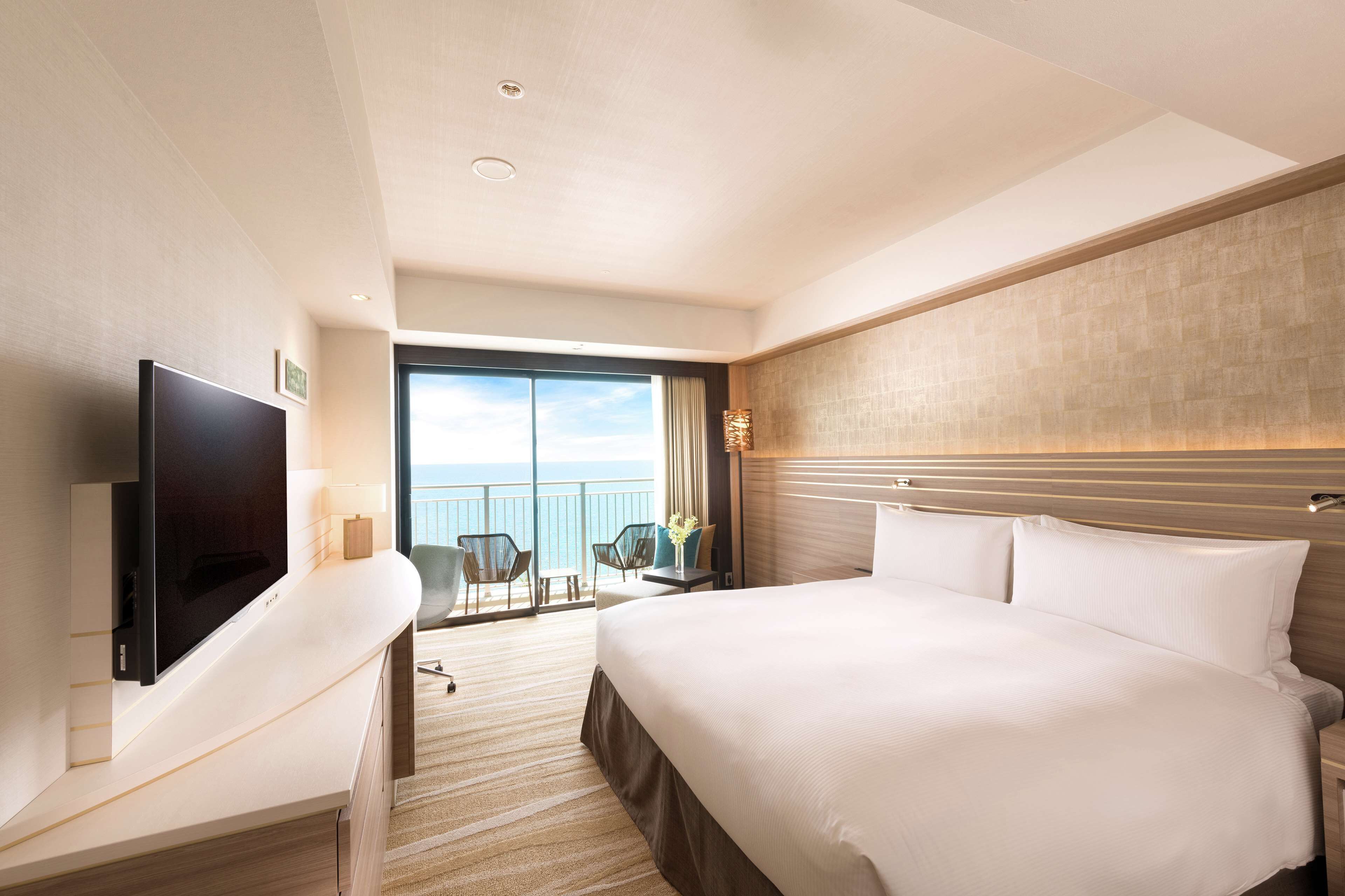 A hotel room with a large bed, a flat-screen TV, a desk with a chair, and a balcony with a view of the ocean.