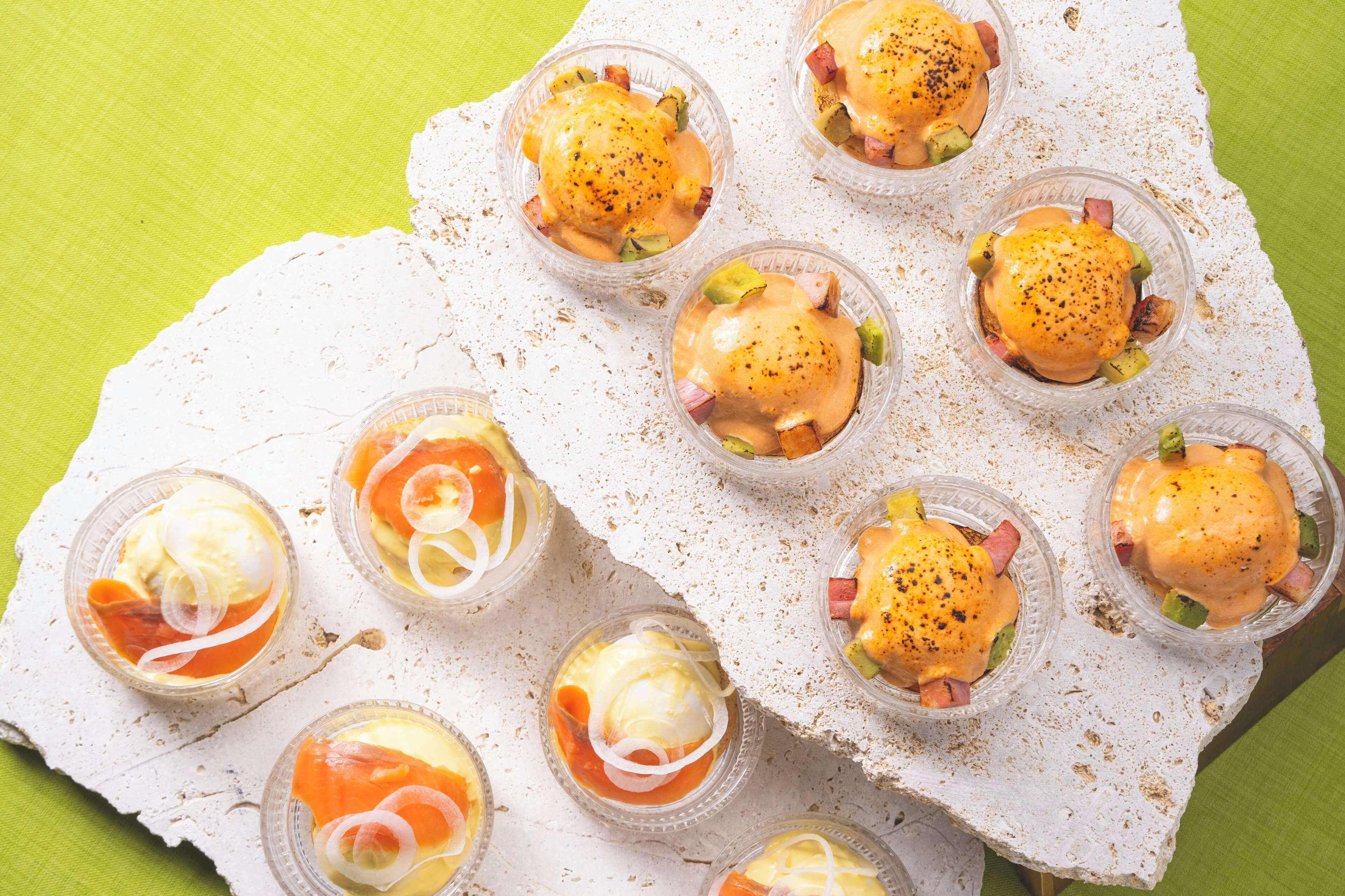 A variety of breakfast dishes in clear cups, including eggs Benedict and omelets, arranged on a white textured surface.