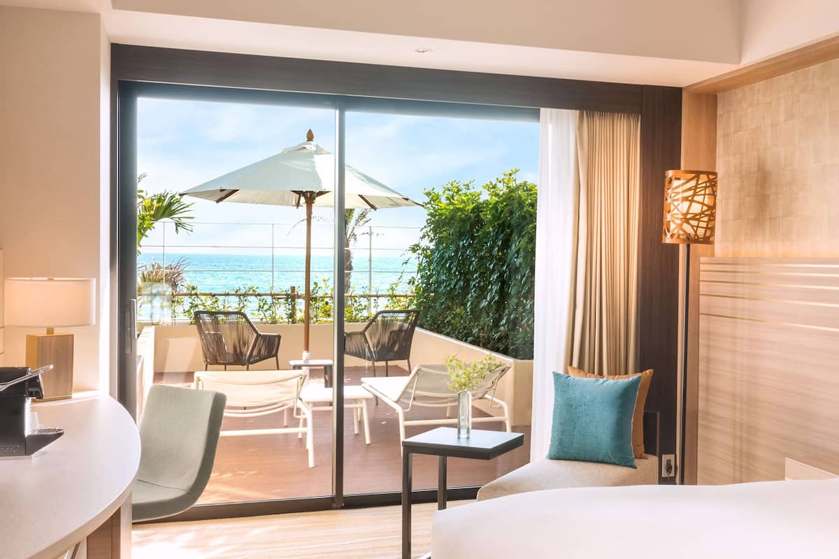 A hotel room with a large sliding glass door leading to a balcony with outdoor seating and a view of the ocean.