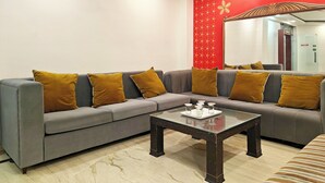 A modern living room with a grey sectional sofa adorned with mustard pillows, a glass coffee table, and a red wall with gold stars and a butterfly design. - Hotel Apple Villa @ Paharganj (New Delhi)