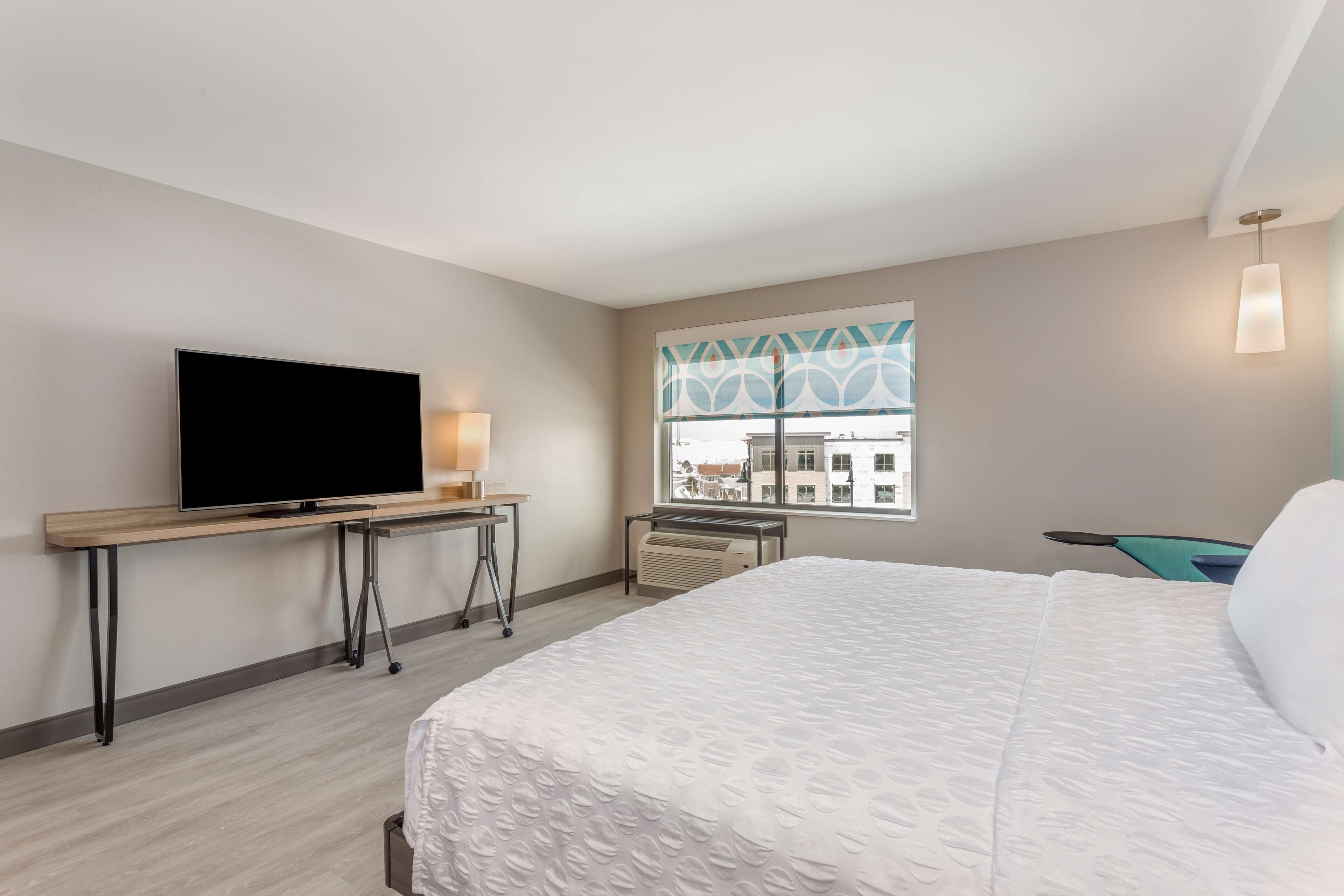 Une chambre d’hôtel avec un lit, un téléviseur posé sur un bureau, une fenêtre donnant sur des immeubles et une lampe.