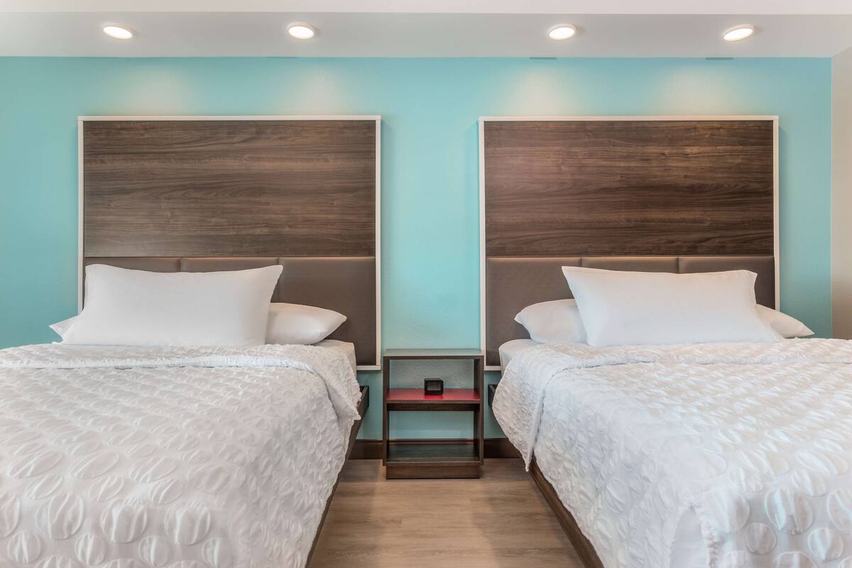 Une chambre d’hôtel avec deux lits, chacun doté d’une tête de lit en bois, de draps blancs et d’une petite table de chevet placée entre les deux.