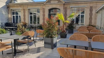 Ein Terrassenbereich im Freien mit Tischen und Stühlen, umgeben von einem Gebäude mit großen Fenstern und Zierelementen.