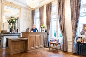 Un hall d’hôtel avec un comptoir d’accueil en bois, un grand miroir et des rideaux à motifs.