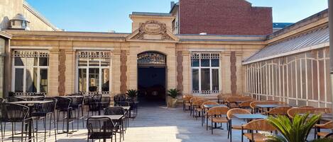 An outdoor cafe area with tables and chairs, a central entrance, and a building facade with a sign.