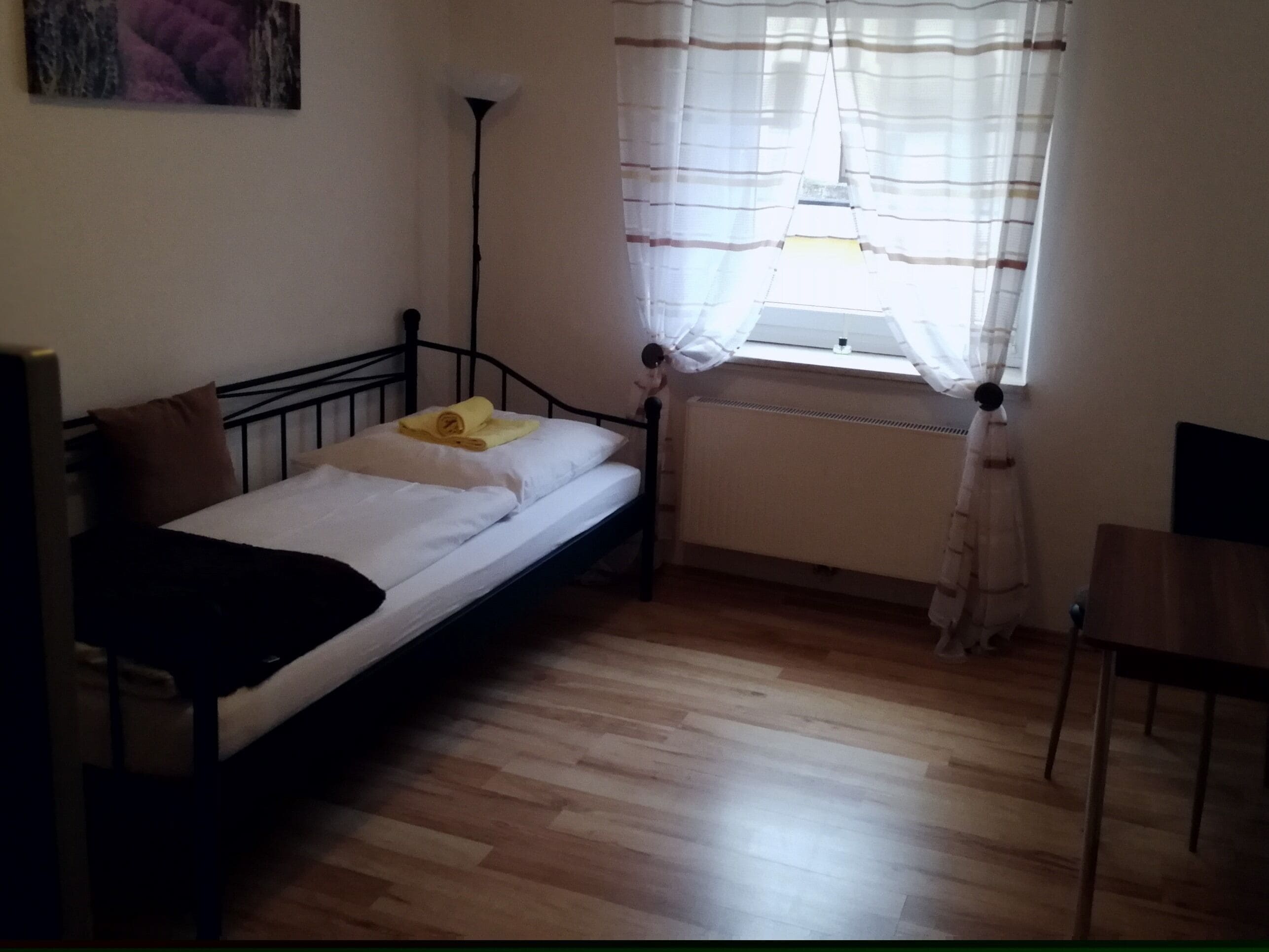 Ein Einzelbett mit weißer Matratze und braunem Kopfkissen, ein kleiner Tisch, eine Stehlampe und ein Fenster mit durchsichtigen Vorhängen.
