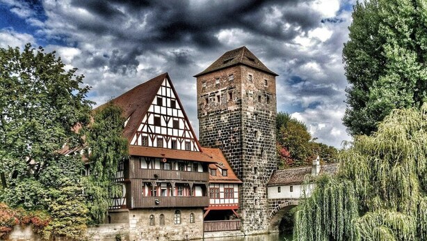 A traditional half-timbered building with a tower, reflecting in a calm river, surrounded by lush greenery. - Sultanias Melanchthon (Nuremberg)