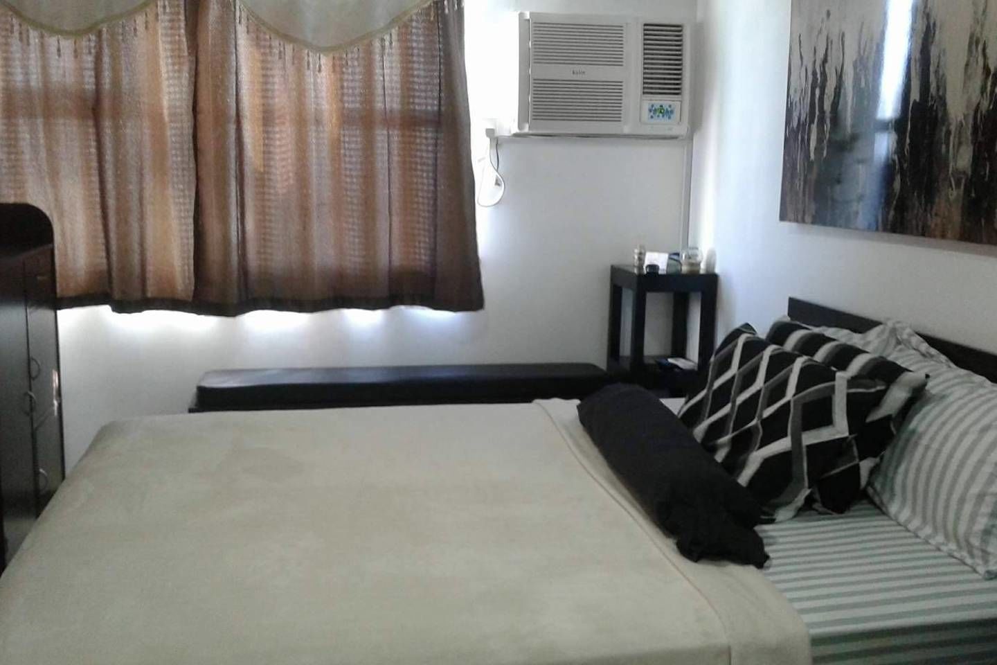 A bedroom with a bed, a bedside table, a wall-mounted air conditioner, and a window with curtains.