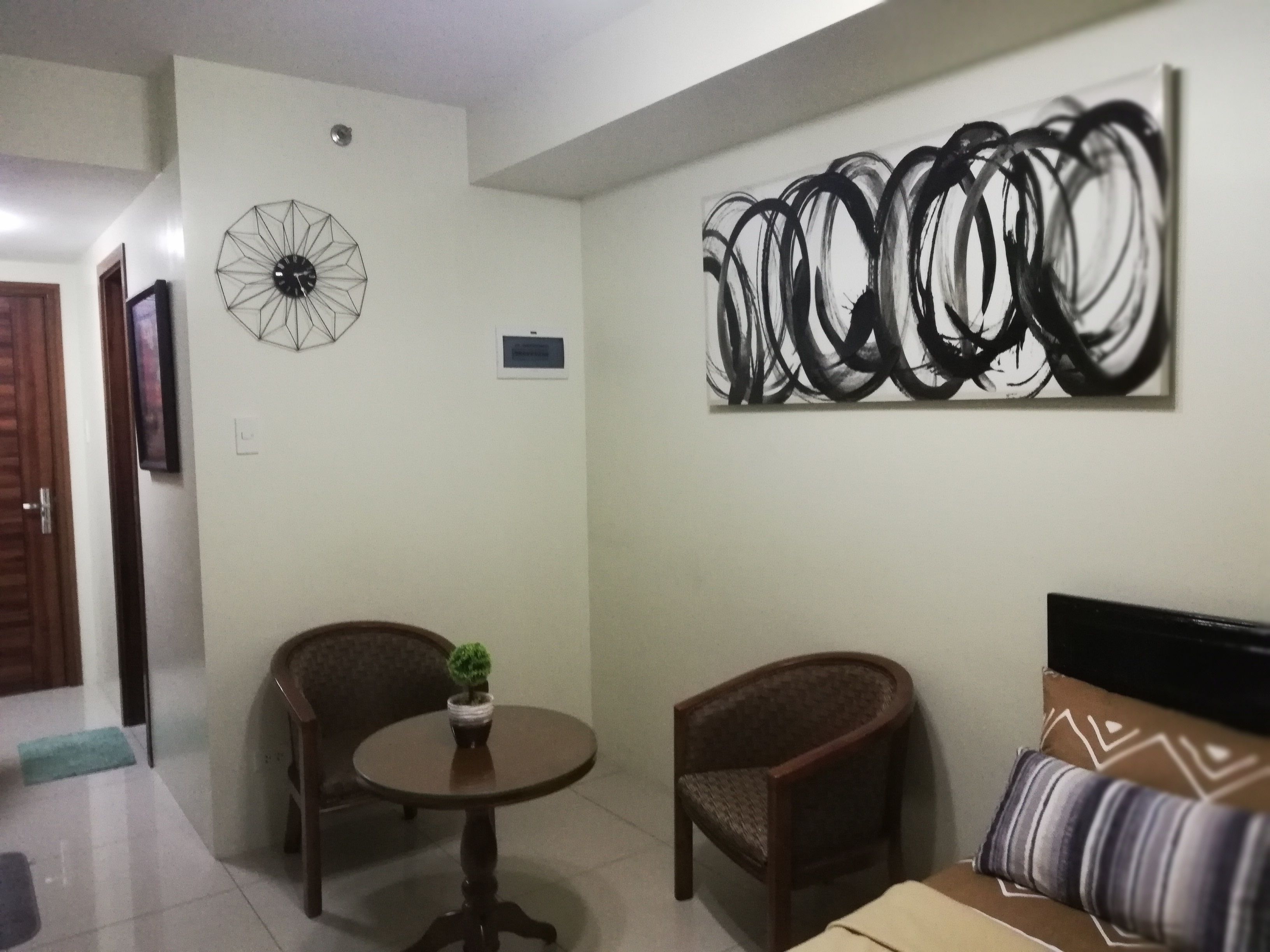 Una sala de estar con una mesa redonda, dos sillas de mimbre, un sofá con cojines estampados, un reloj de pared y un cuadro abstracto grande.