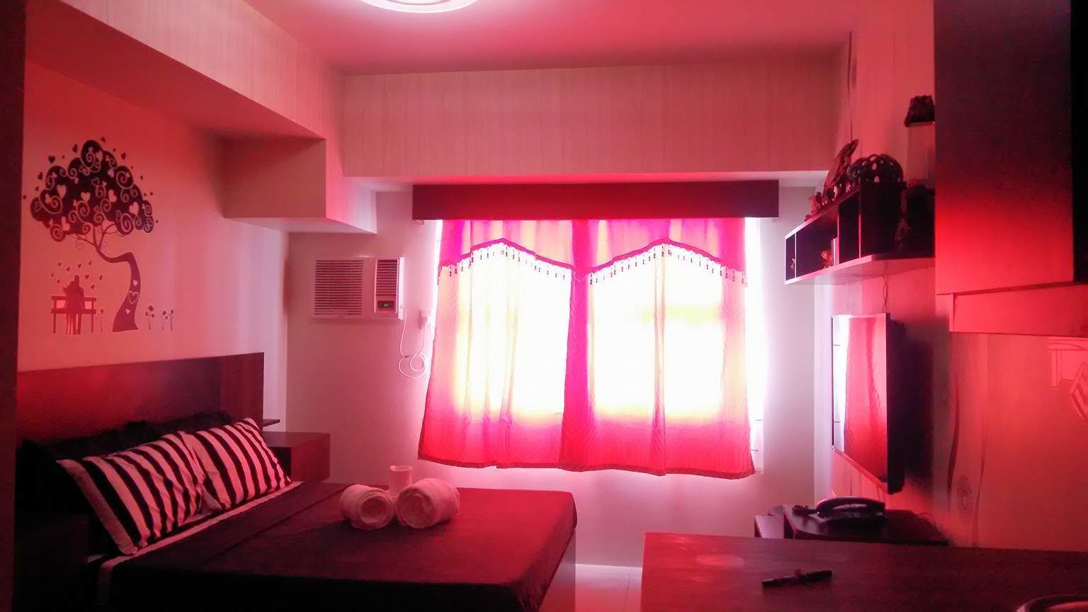 Een kamer met een bed, een tafel, een televisie en een raam met rode gordijnen.