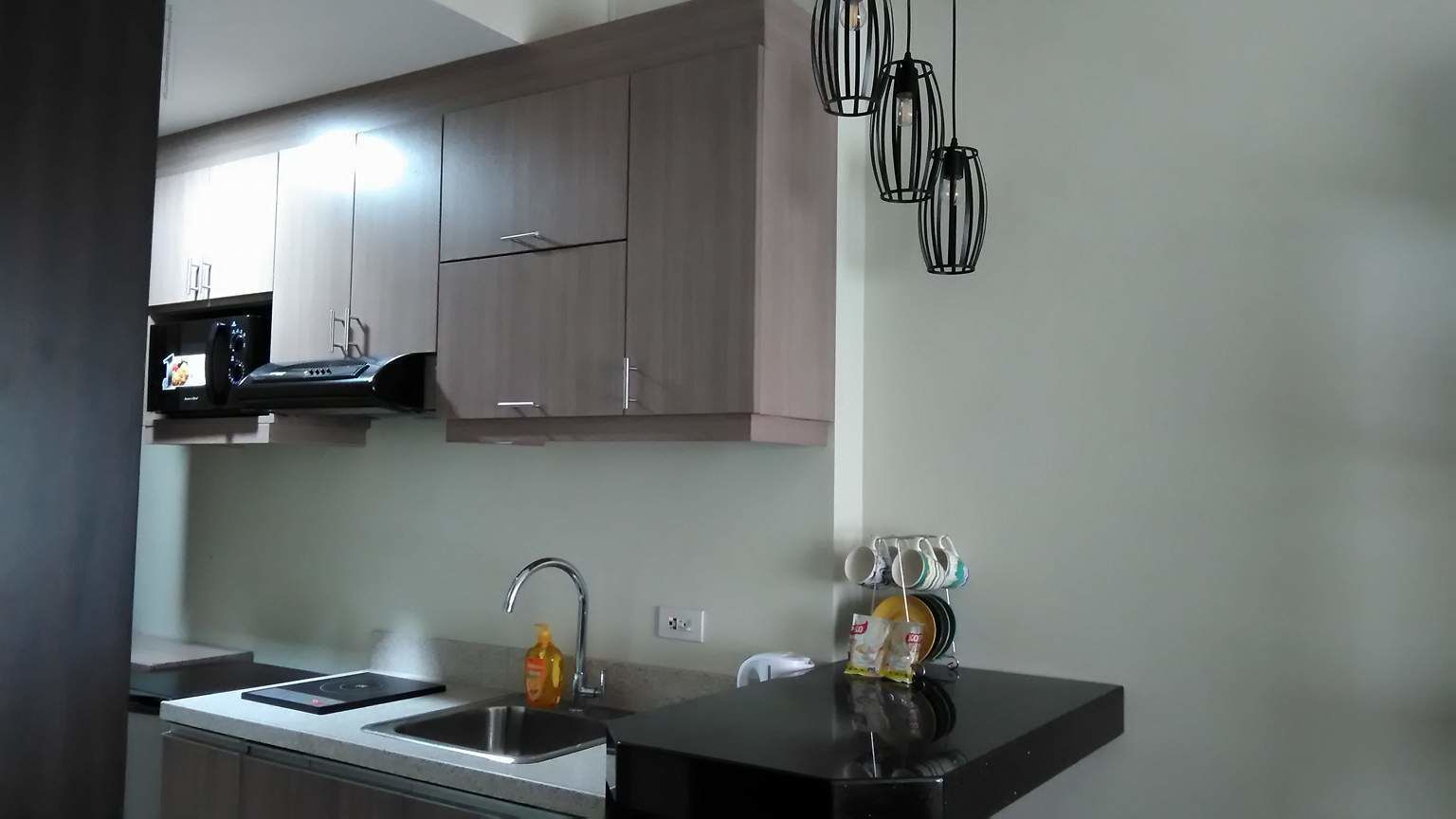 A modern kitchen with a built-in microwave, stainless steel sink, and a black countertop. There are hanging pendant lights above the sink and a small shelf with kitchen items.