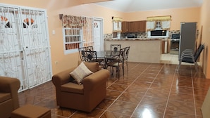 A living room with a brown sofa and a dining table with chairs. There is a kitchen area with wooden cabinets and a refrigerator. - Ri'Biero's Holiday Apartments (Crown Point)