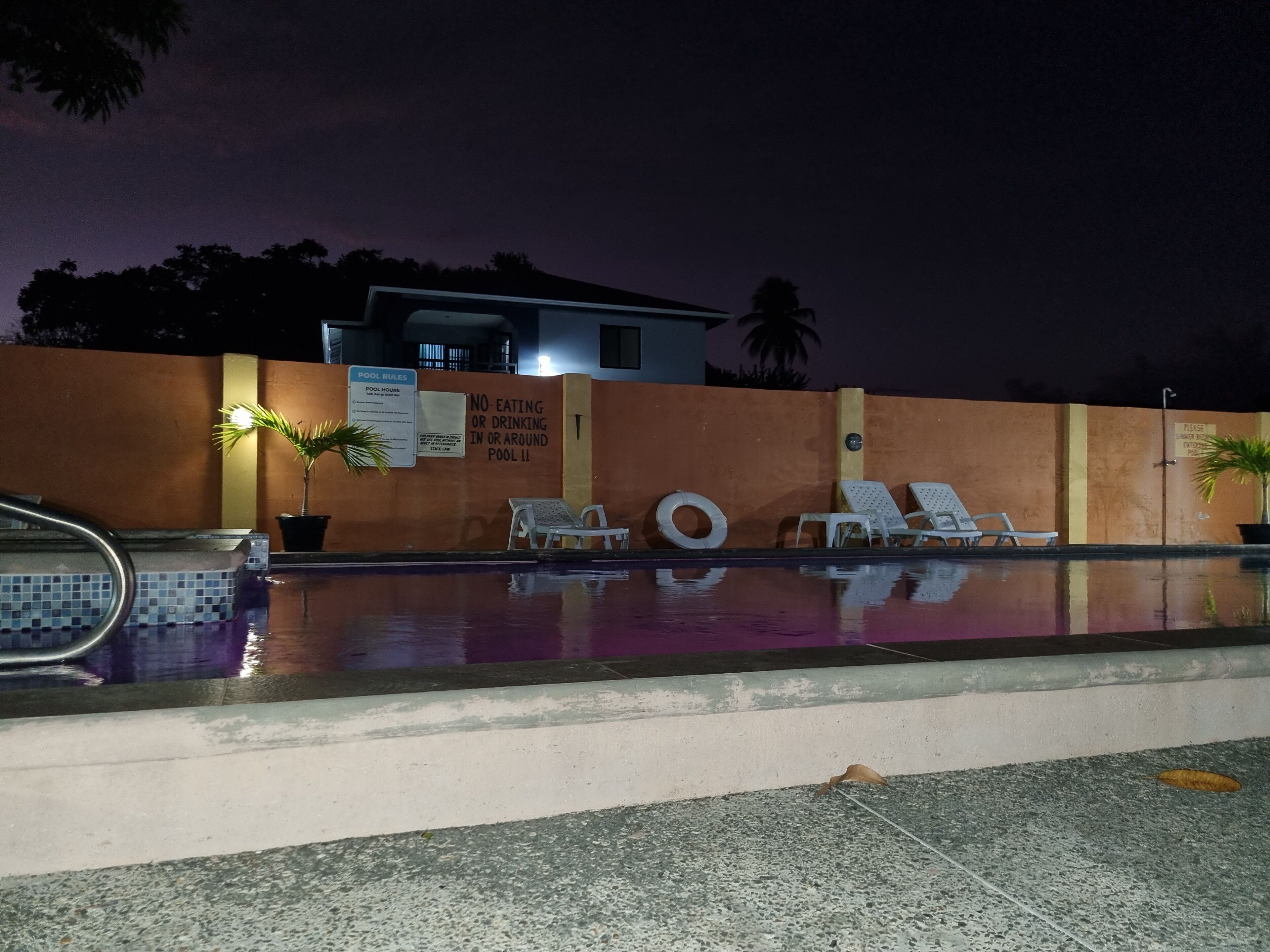A nighttime pool area with lounge chairs, a pool with a tiled edge, and a wall with a 'No-eating or drinking in or around pool' sign.