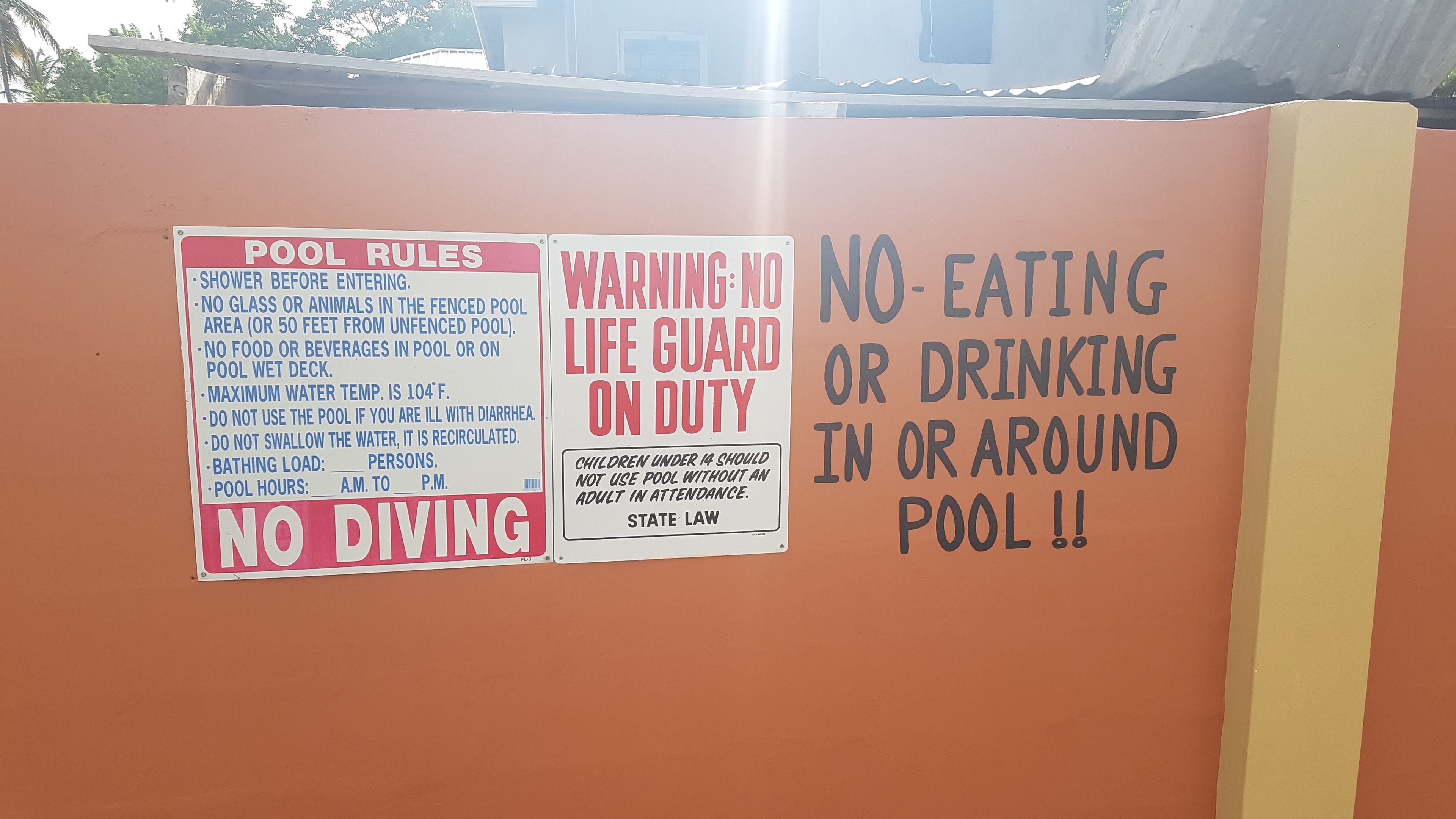 A wall with multiple signs including 'POOL RULES', 'WARNING: NO LIFE GUARD ON DUTY', 'NO EATING OR DRINKING IN OR AROUND POOL !!', and 'NO DIVING'.