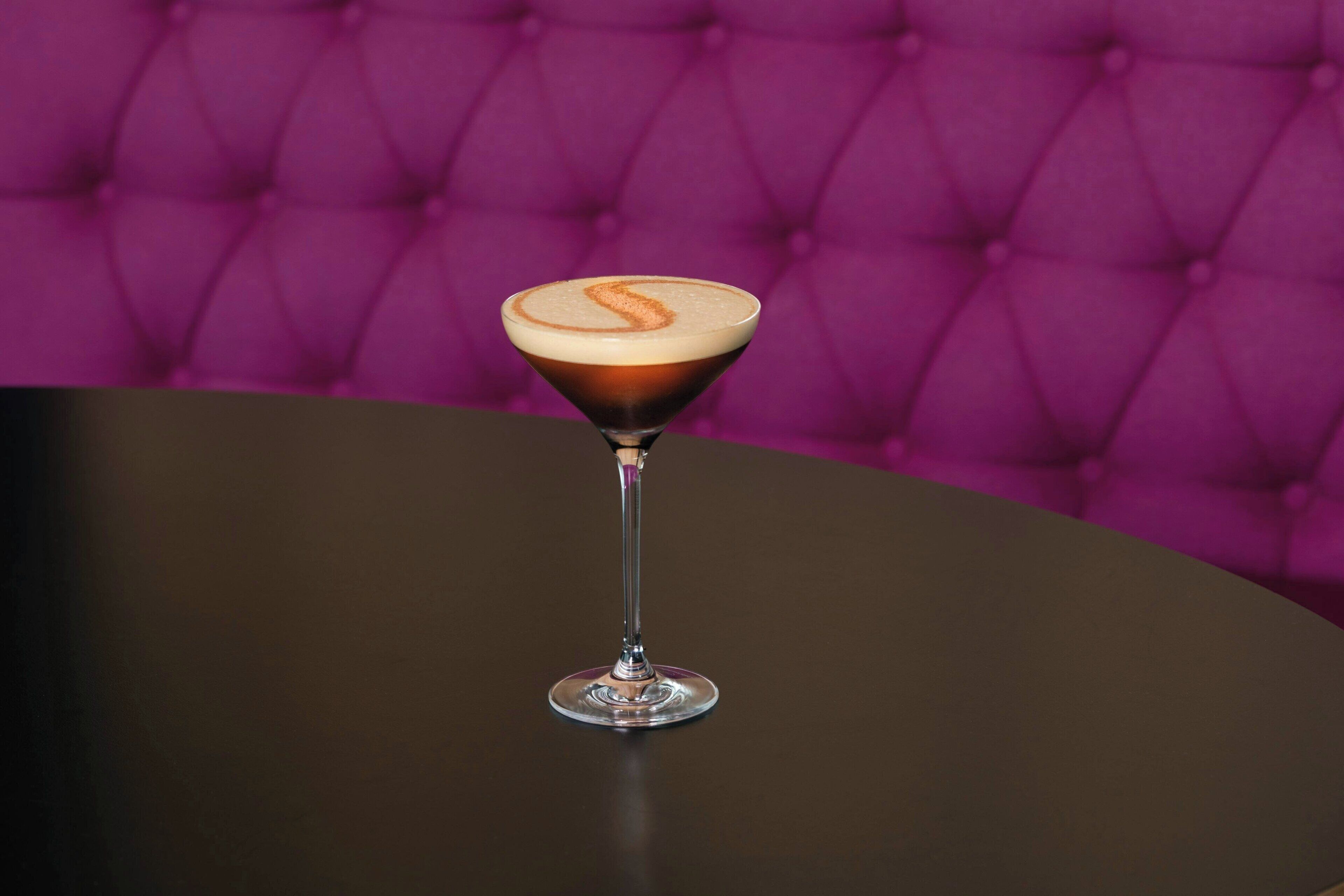 Una copa de martini con una bebida en capas sobre una mesa redonda, frente a un fondo tapizado morado.