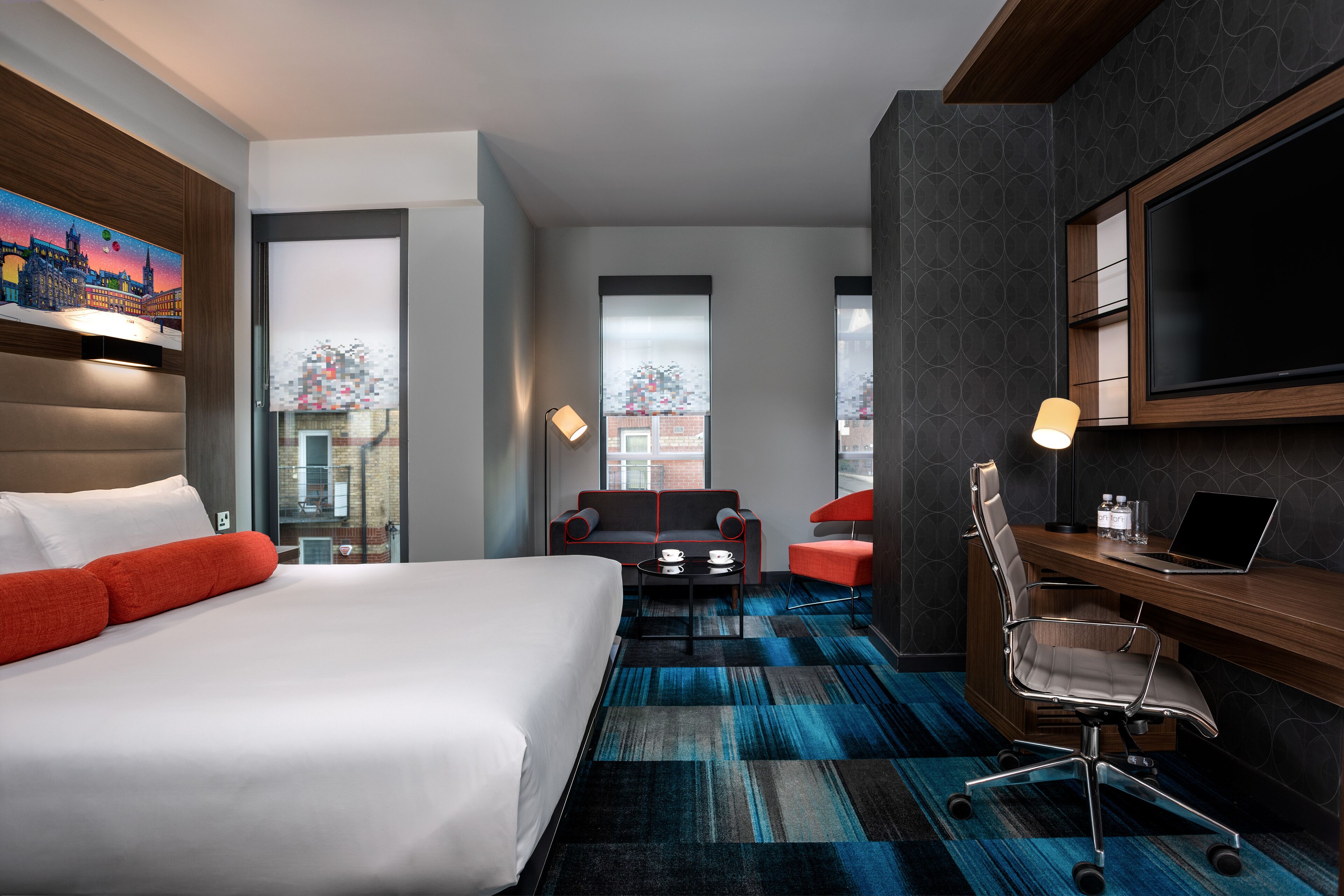 A modern hotel room with a large bed, a desk with a laptop, a chair, a sofa, and a TV.